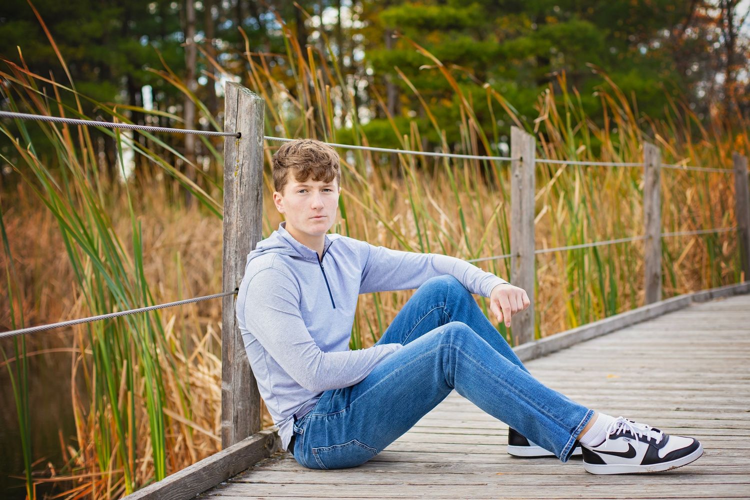 Teen boy in blue hoodie and jeans sitting relaxed on a wooden boardwalk near tall grass outdoors.