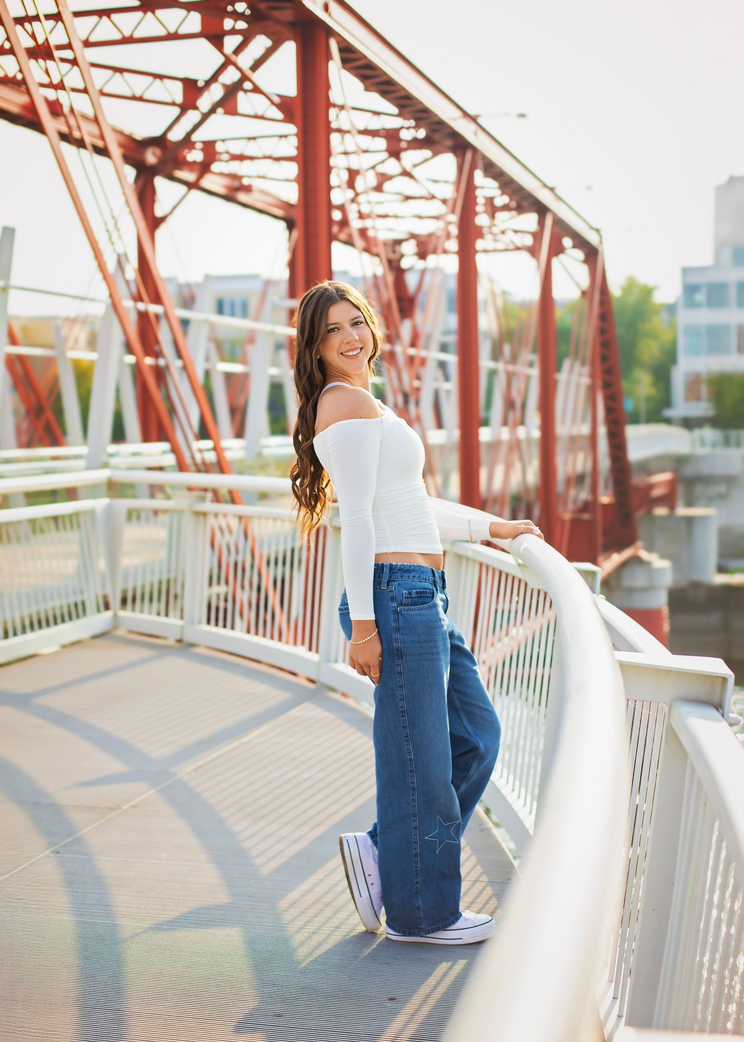 Smiling young woman in white top and jeans poses on red iron bridge walkway in sunlight.
