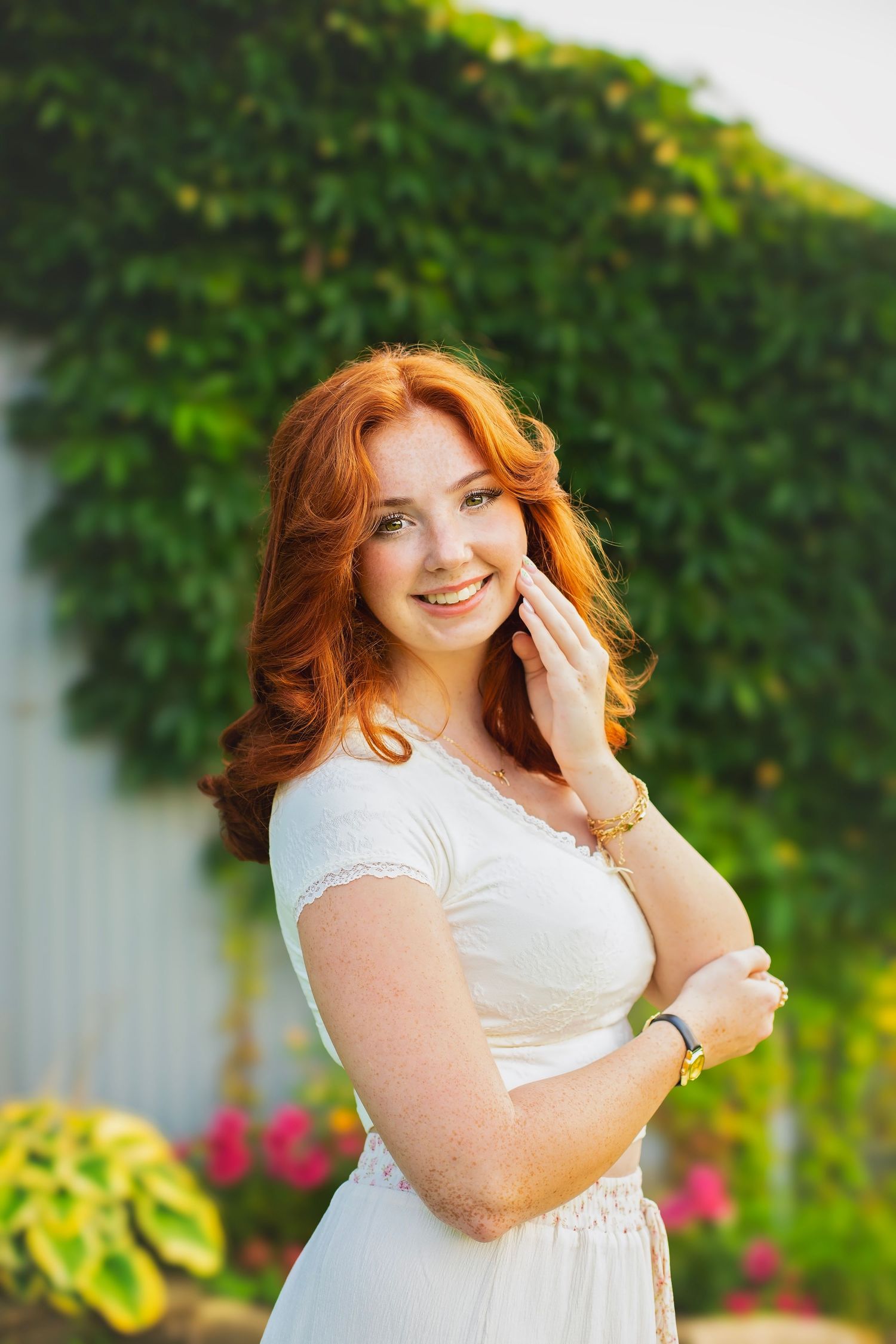 Smiling redhead woman in white dress posing outdoors near ivy-covered wall in warm sunlight.