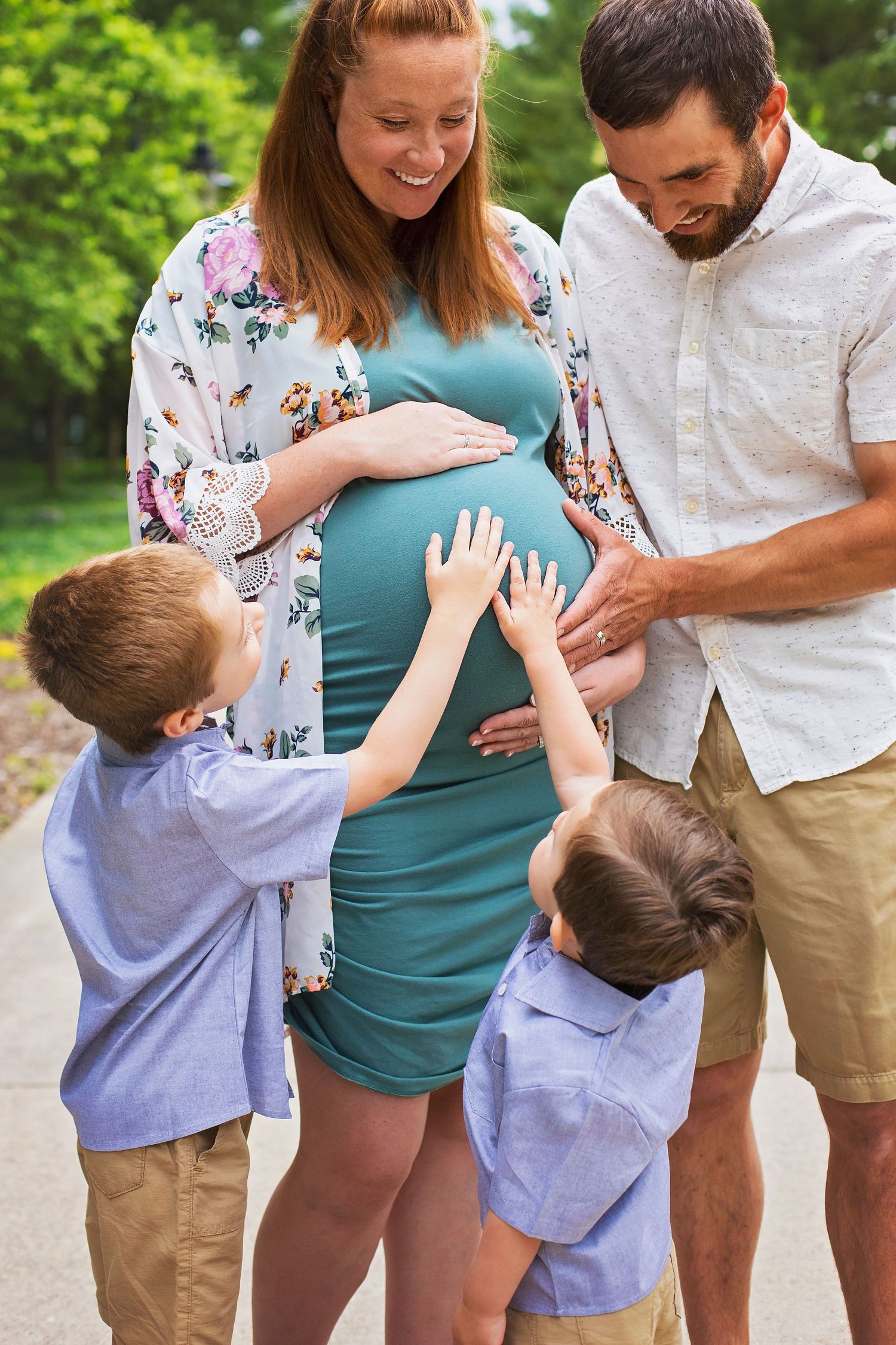 Happy family with two young boys touching pregnant mothers belly outdoors in a green park setting.