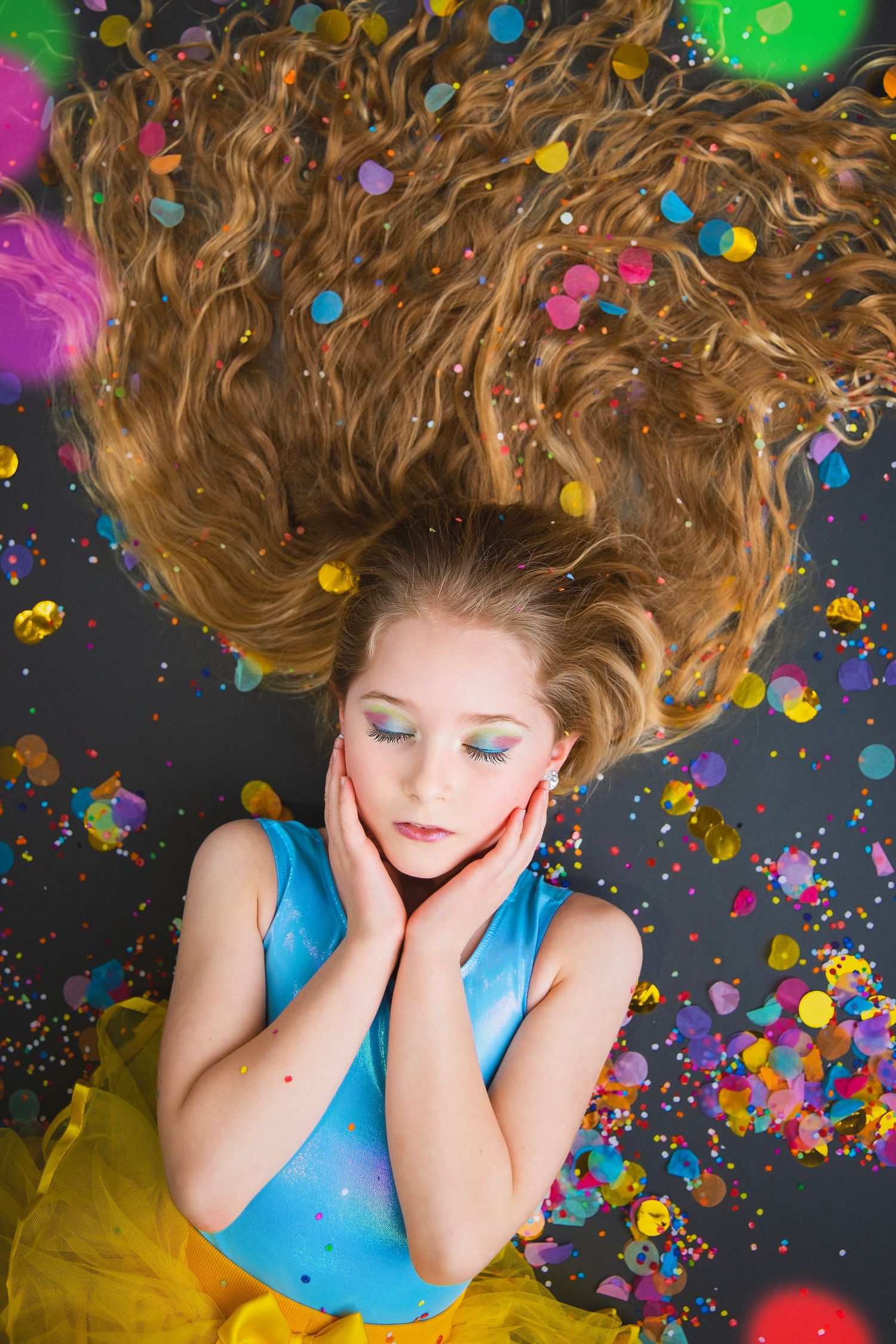A young girl with long curly hair lies on a dark floor with colorful confetti, resting her face in her hands.