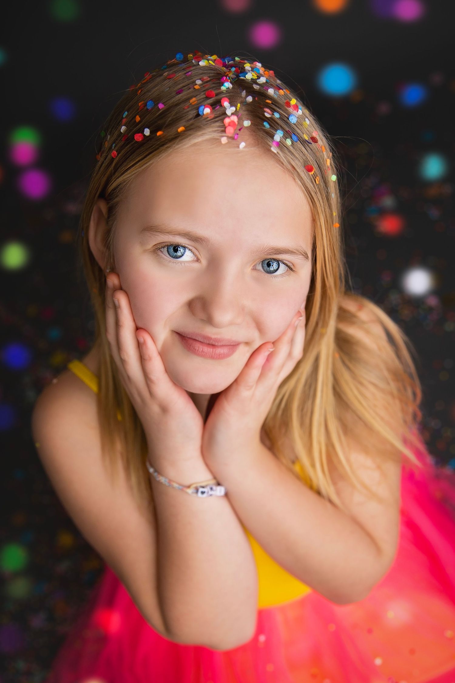 Pretty blonde girl in yellow and pink tutu posing with hands on cheeks amid colorful bokeh.