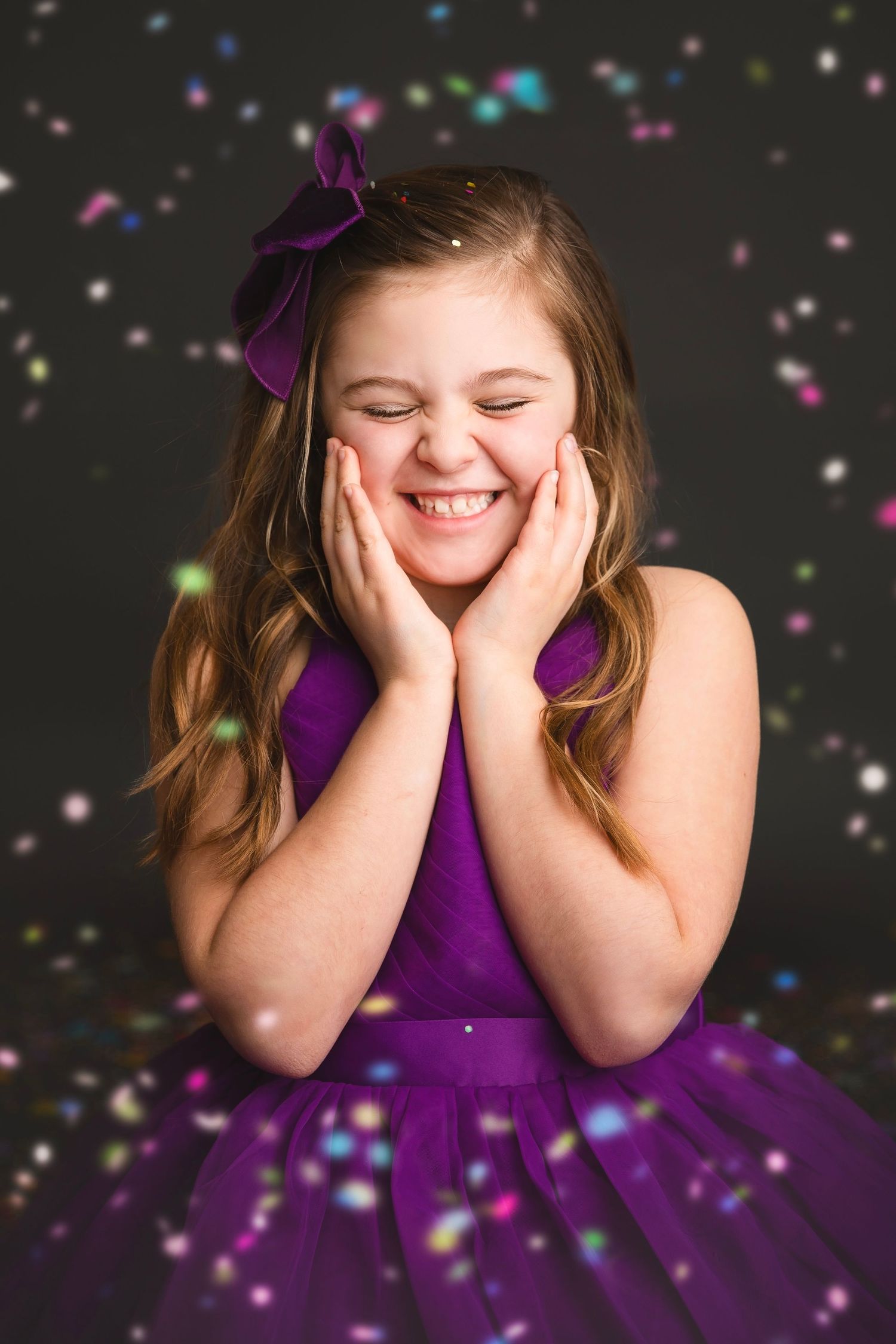 Joyful girl in purple dress with bow, eyes closed, hands on cheeks, with confetti bokeh background.