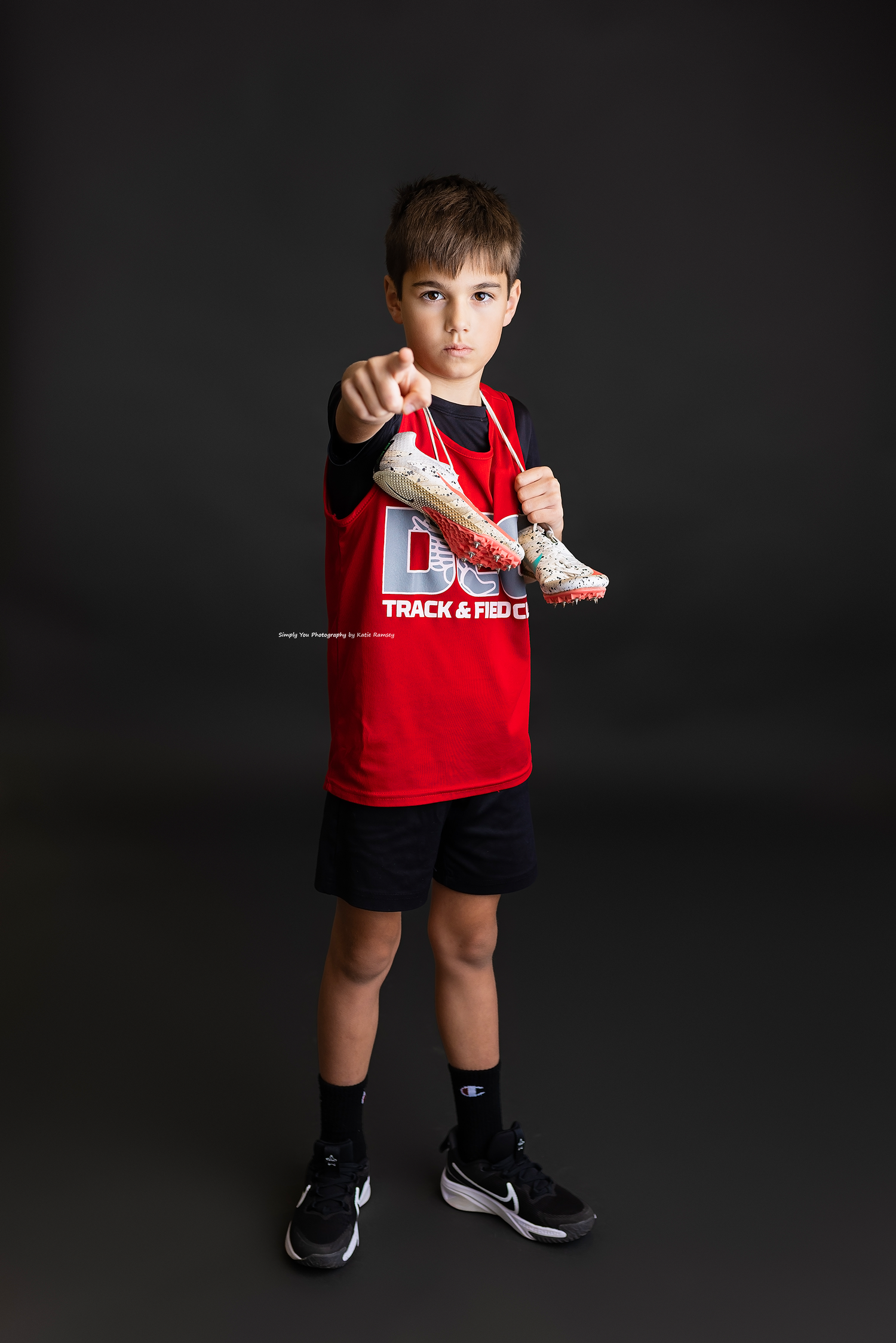 Young boy in red track jersey points forward holding cleats against dark background.