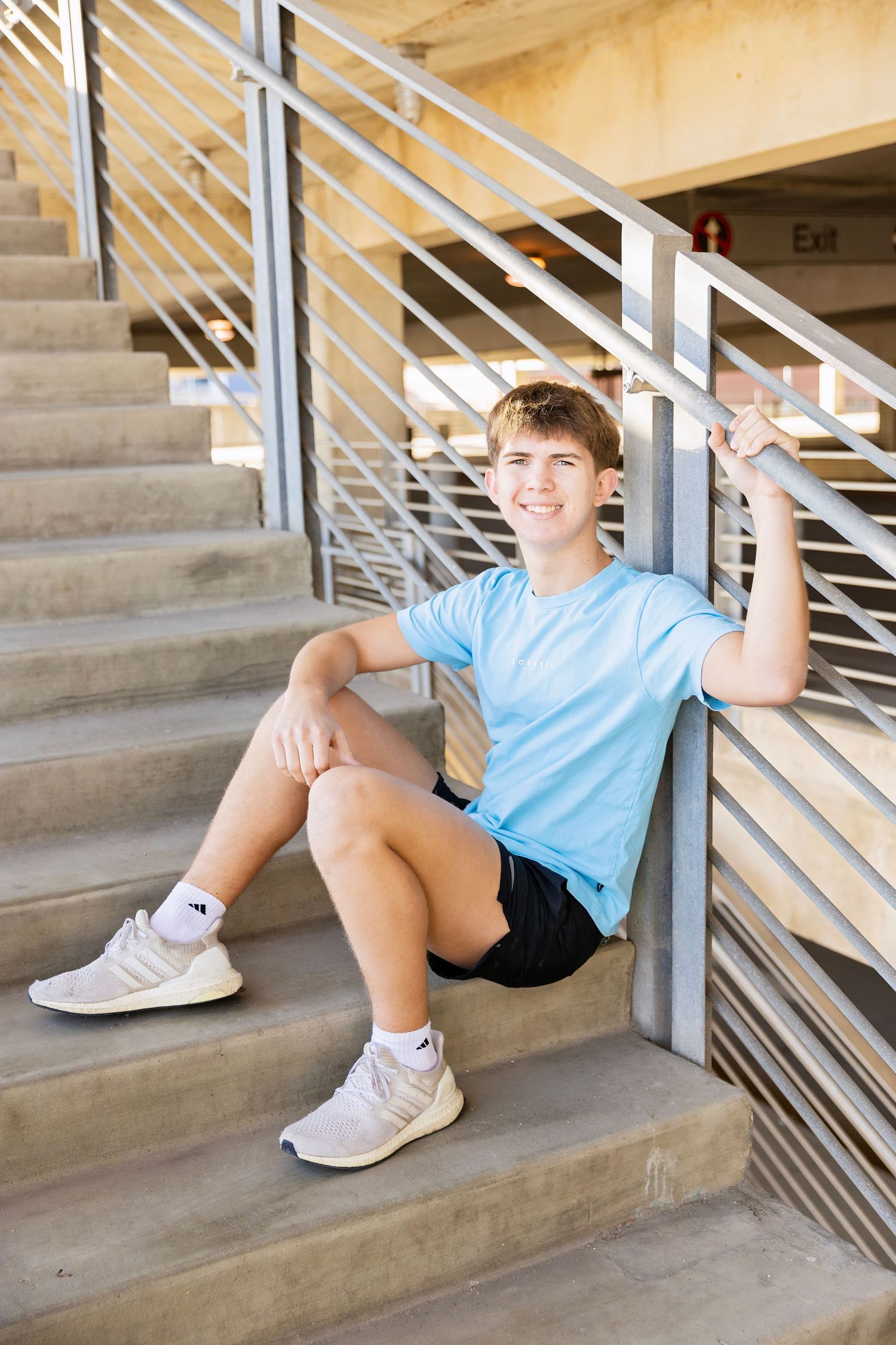 Smiling teen boy in blue shirt sitting on concrete stairs holding metal railing.