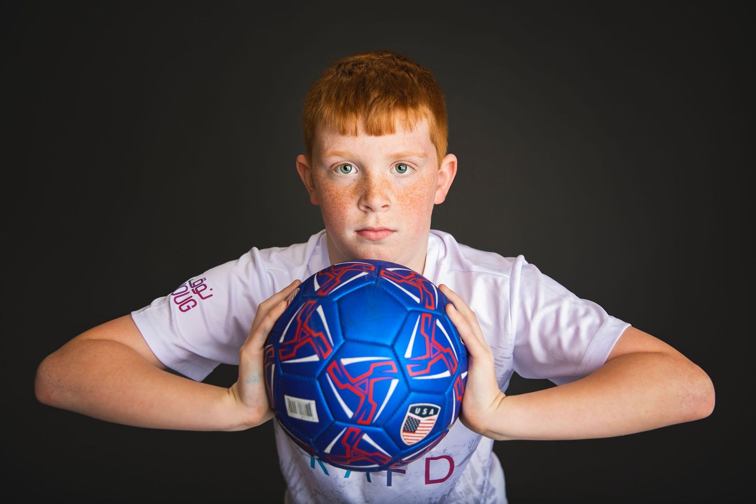 Red-haired boy in white jersey holds blue and red soccer ball toward camera.