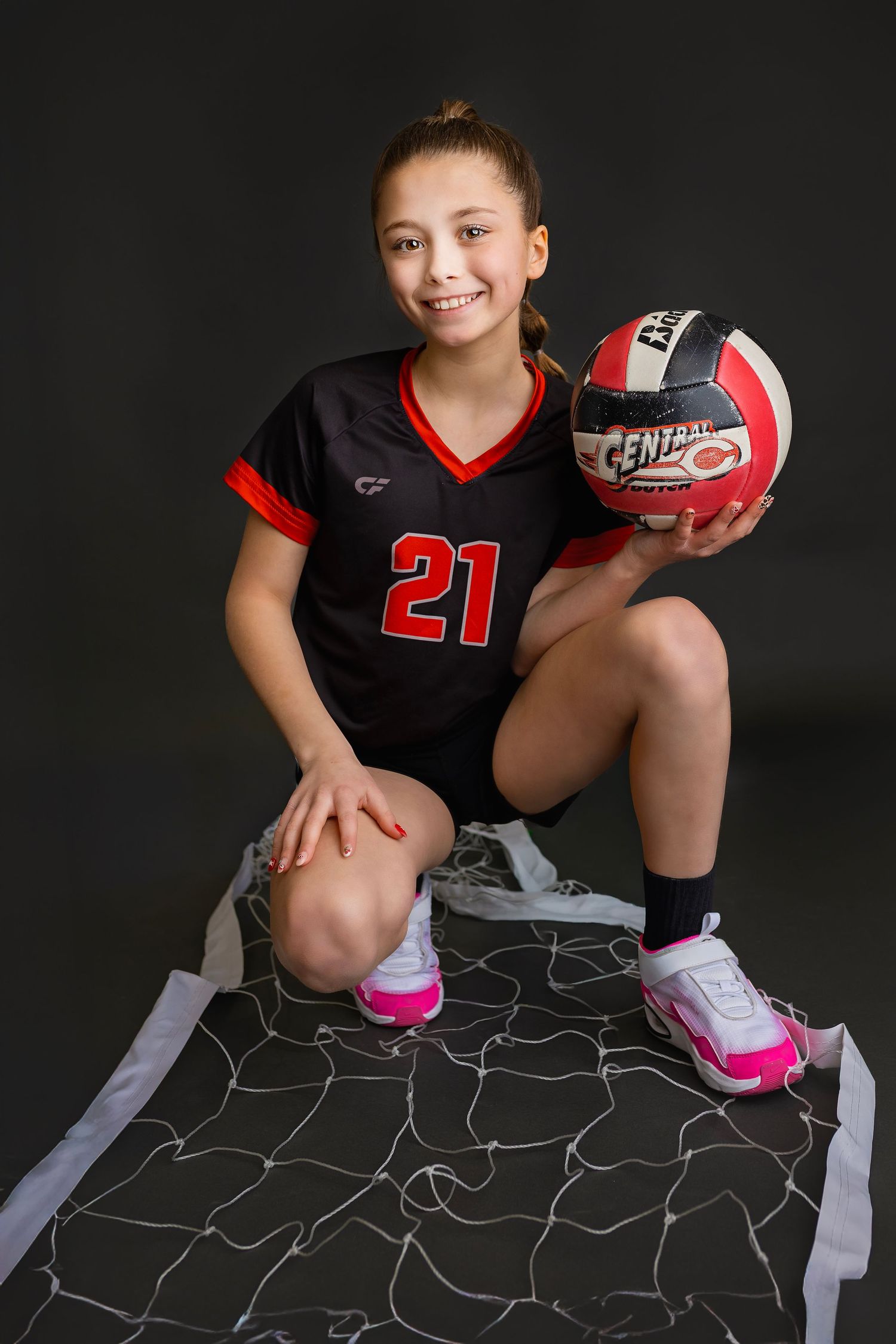 Young girl in number 21 volleyball uniform crouching on net holding Central volleyball smiling at camera.