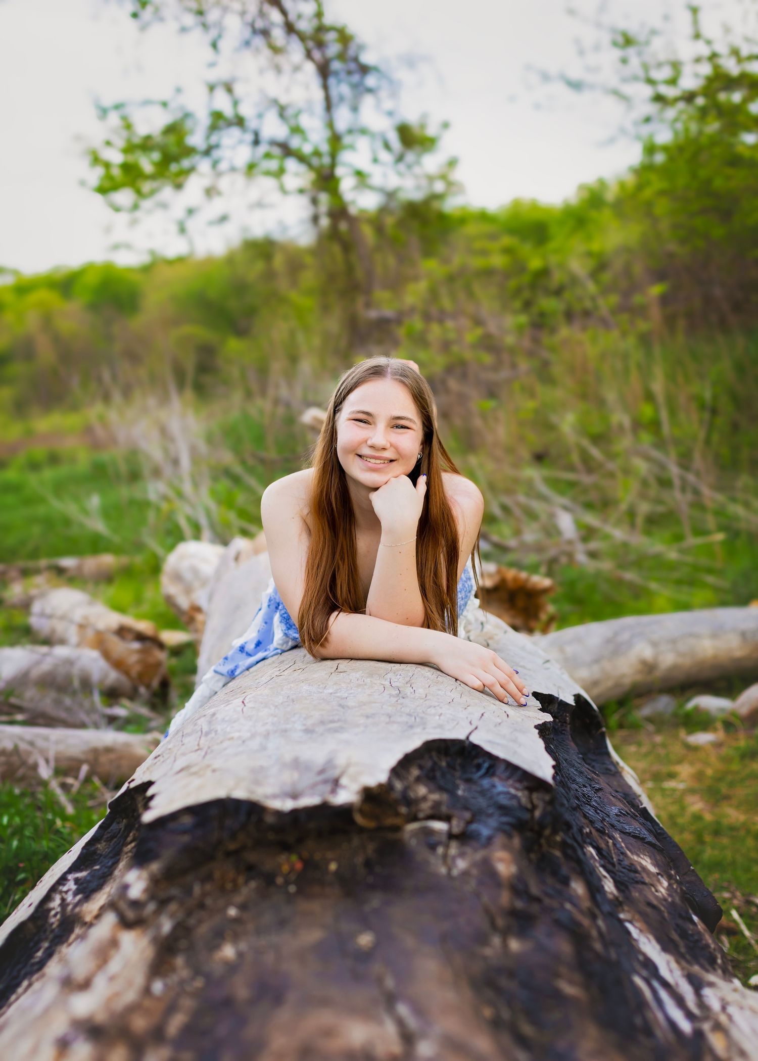 Smiling teenage girl with long brown hair lying on fallen log in green outdoor nature setting.
