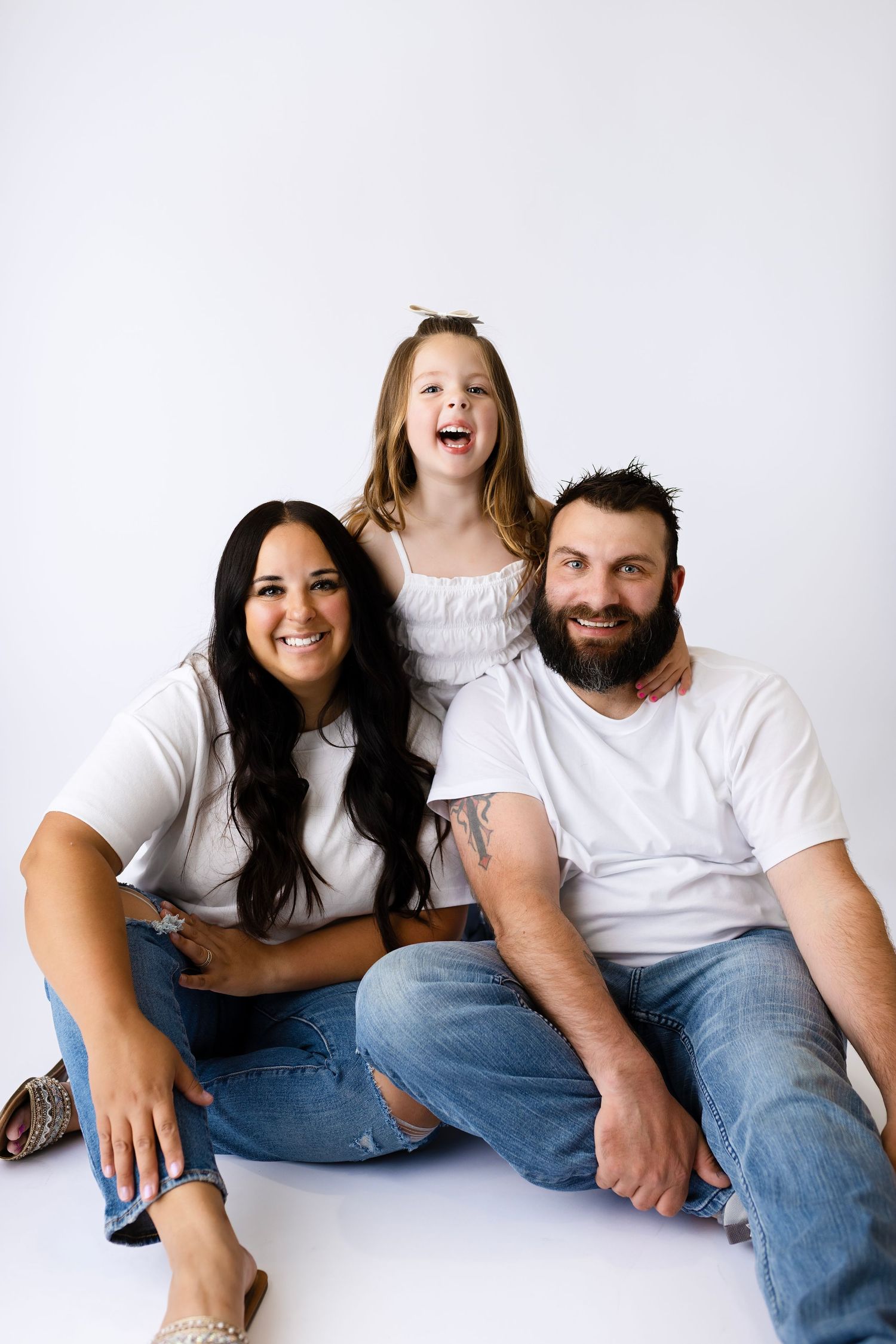 A smiling family of three in white tops and jeans poses together against a white background for a portrait.