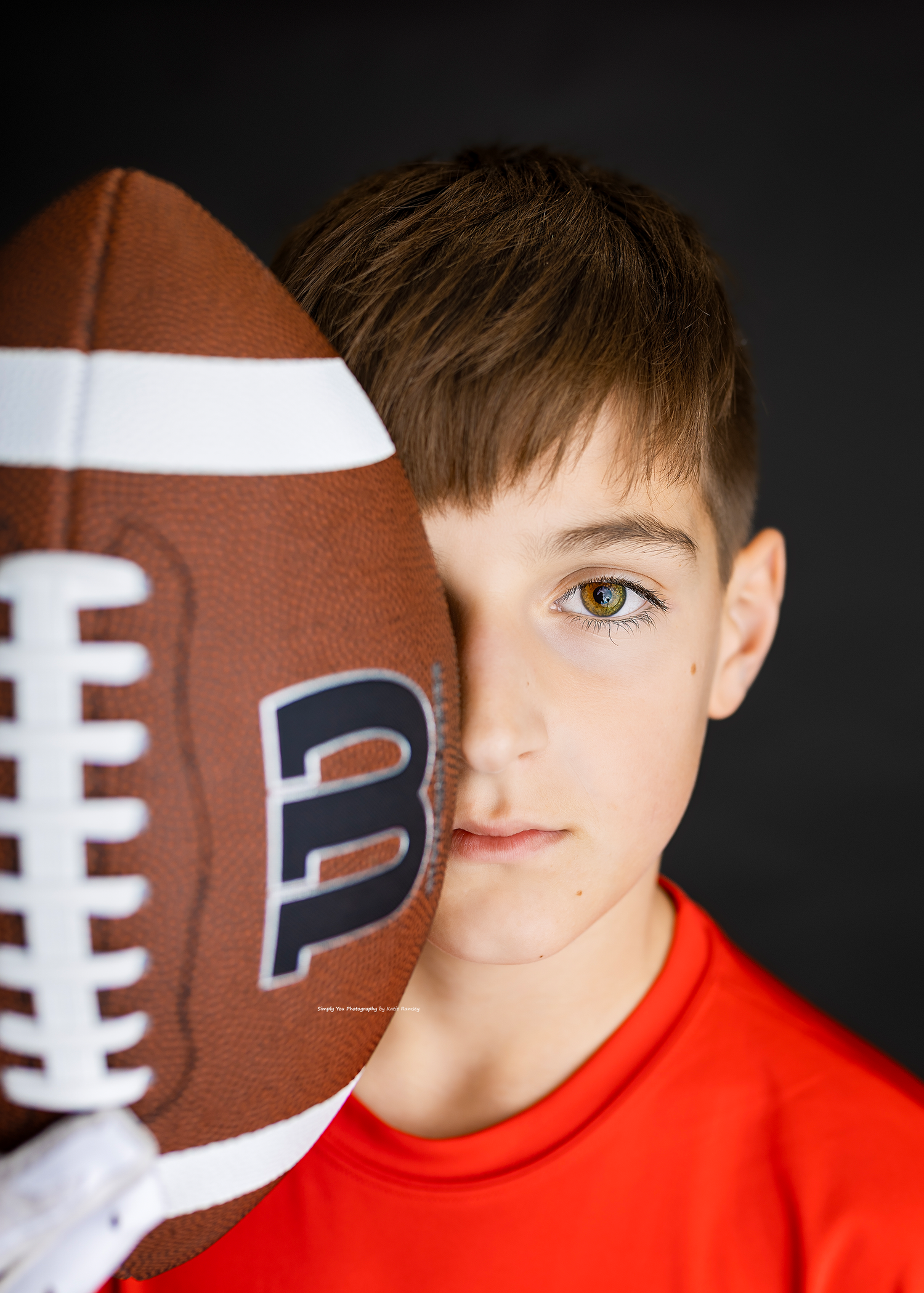 Boy in red shirt holds football covering half his face, showcasing green eyes.