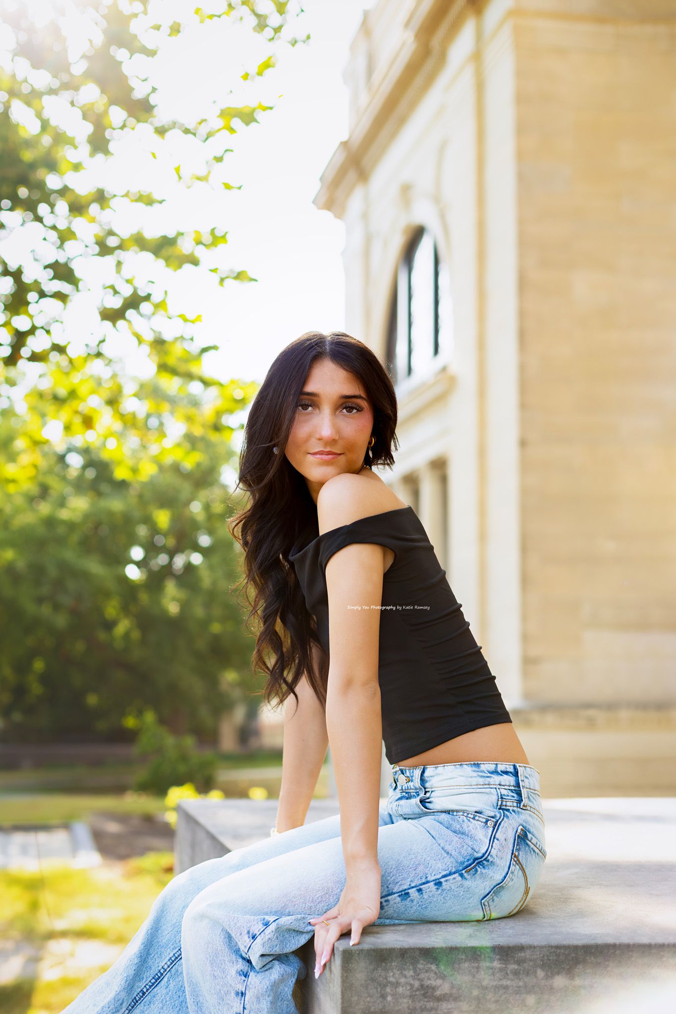 Young woman with dark hair in black off-shoulder top and jeans sitting outdoors near a stone building.