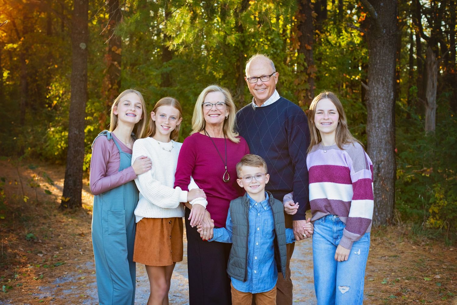 Family of six smiling together on a sunny tree-lined path in autumn.