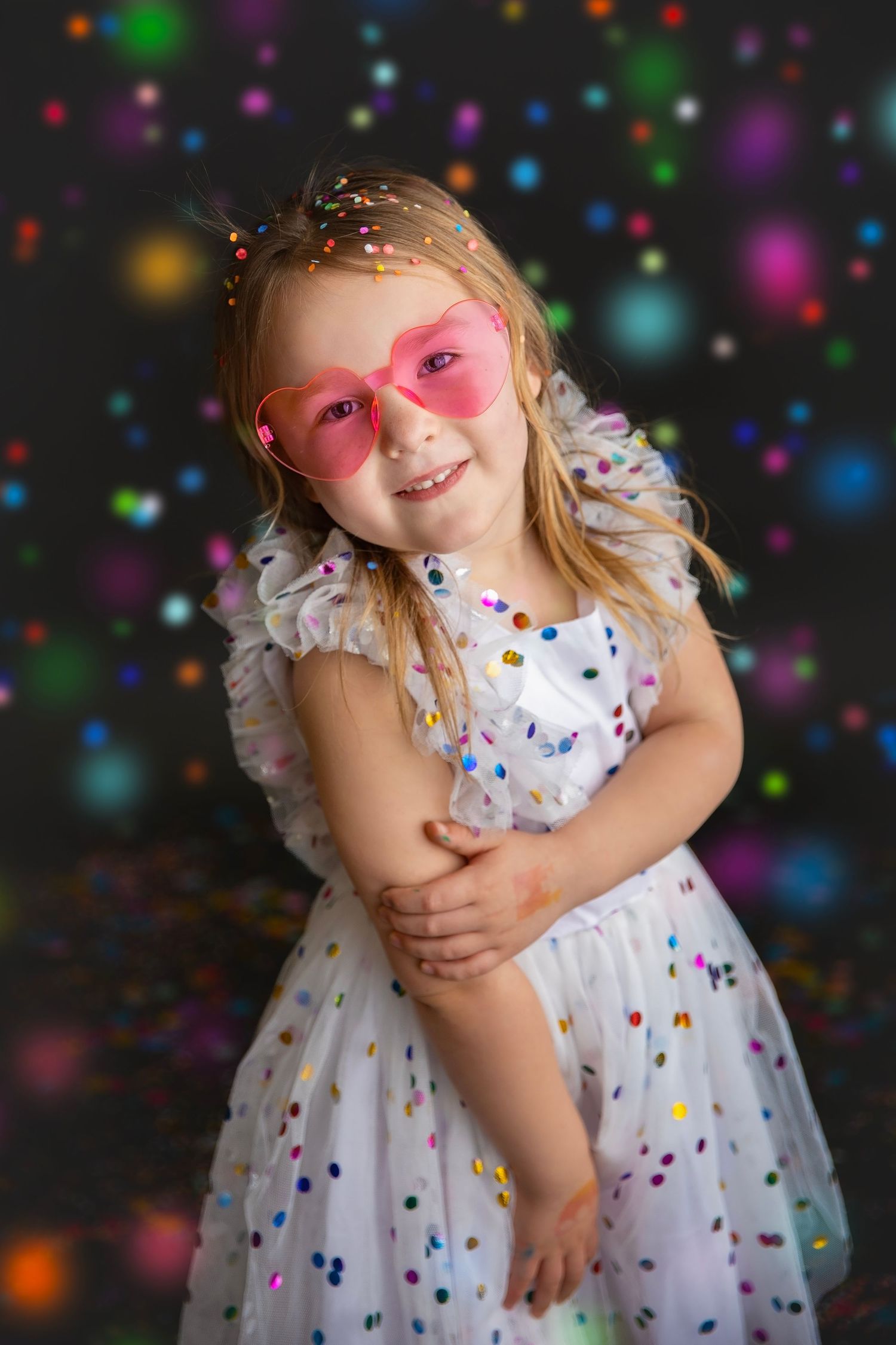 A smiling young girl in a white polka dot dress wears pink heart sunglasses surrounded by colorful bokeh lights.
