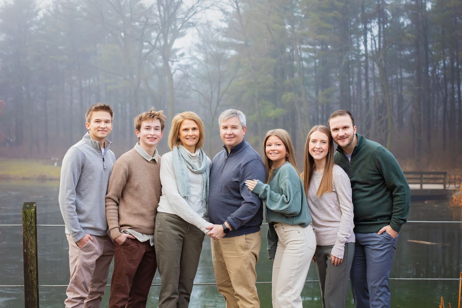 Family of seven posing on a foggy lakeside dock in coordinated earth-tone outfits.