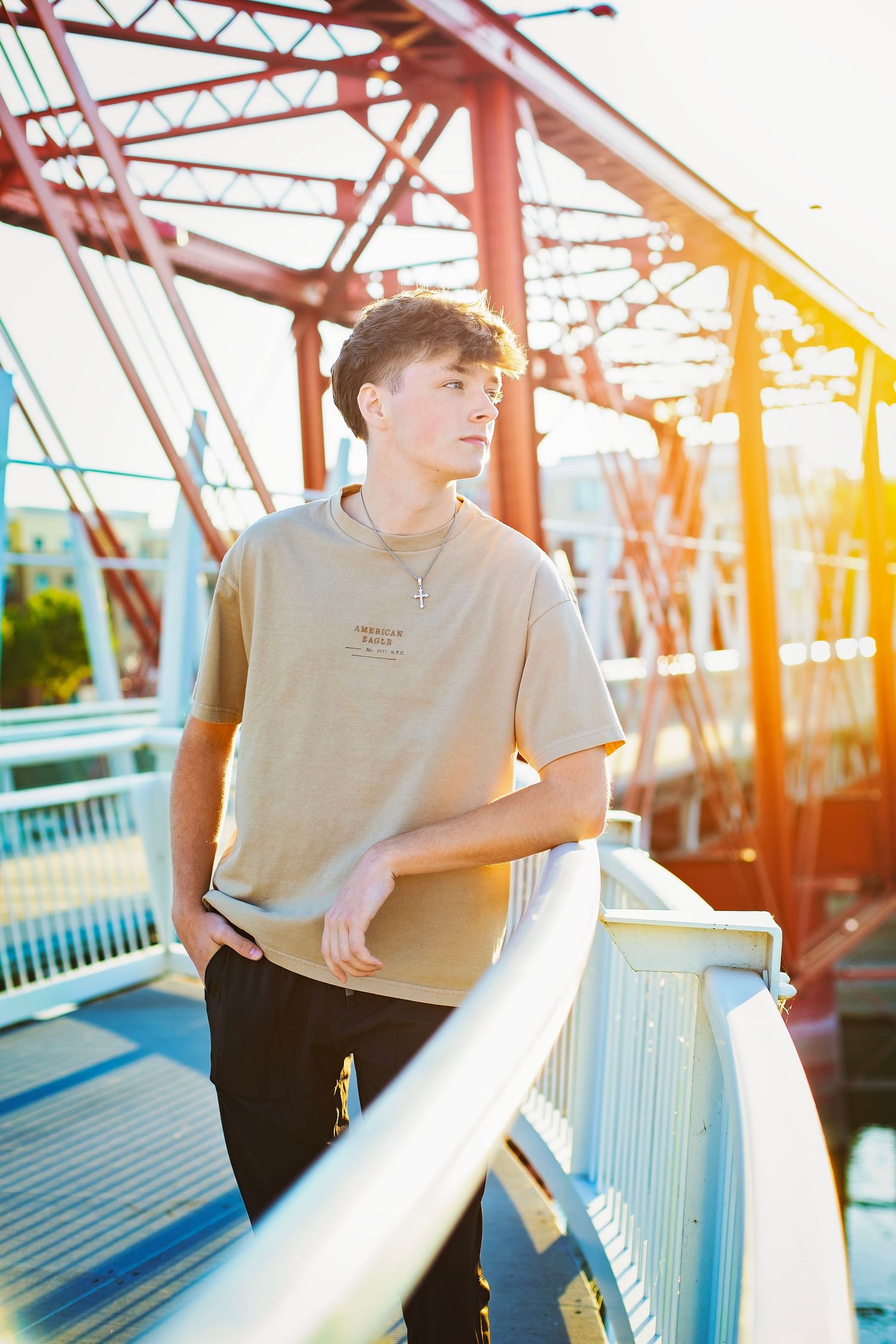 Teen boy in beige t-shirt standing on bridge railing with golden sunset light behind him.