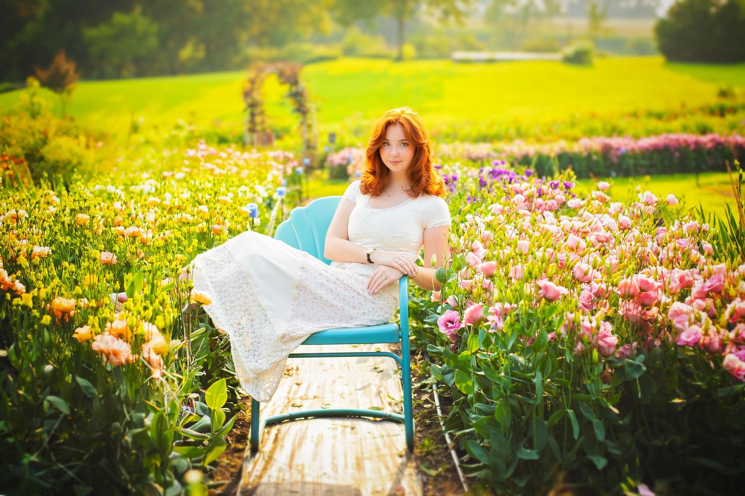 Redhead woman in white dress sitting on blue chair surrounded by colorful tulips in sunny garden.