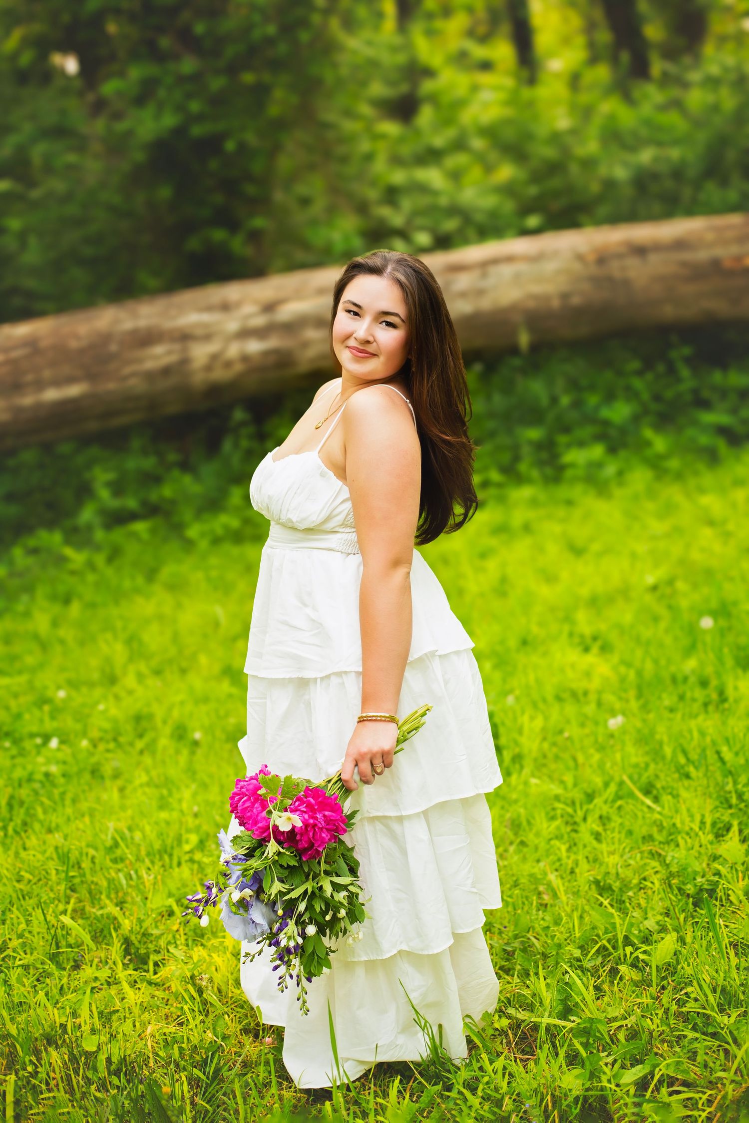 Smiling young woman in white ruffled dress holding pink floral bouquet outdoors.