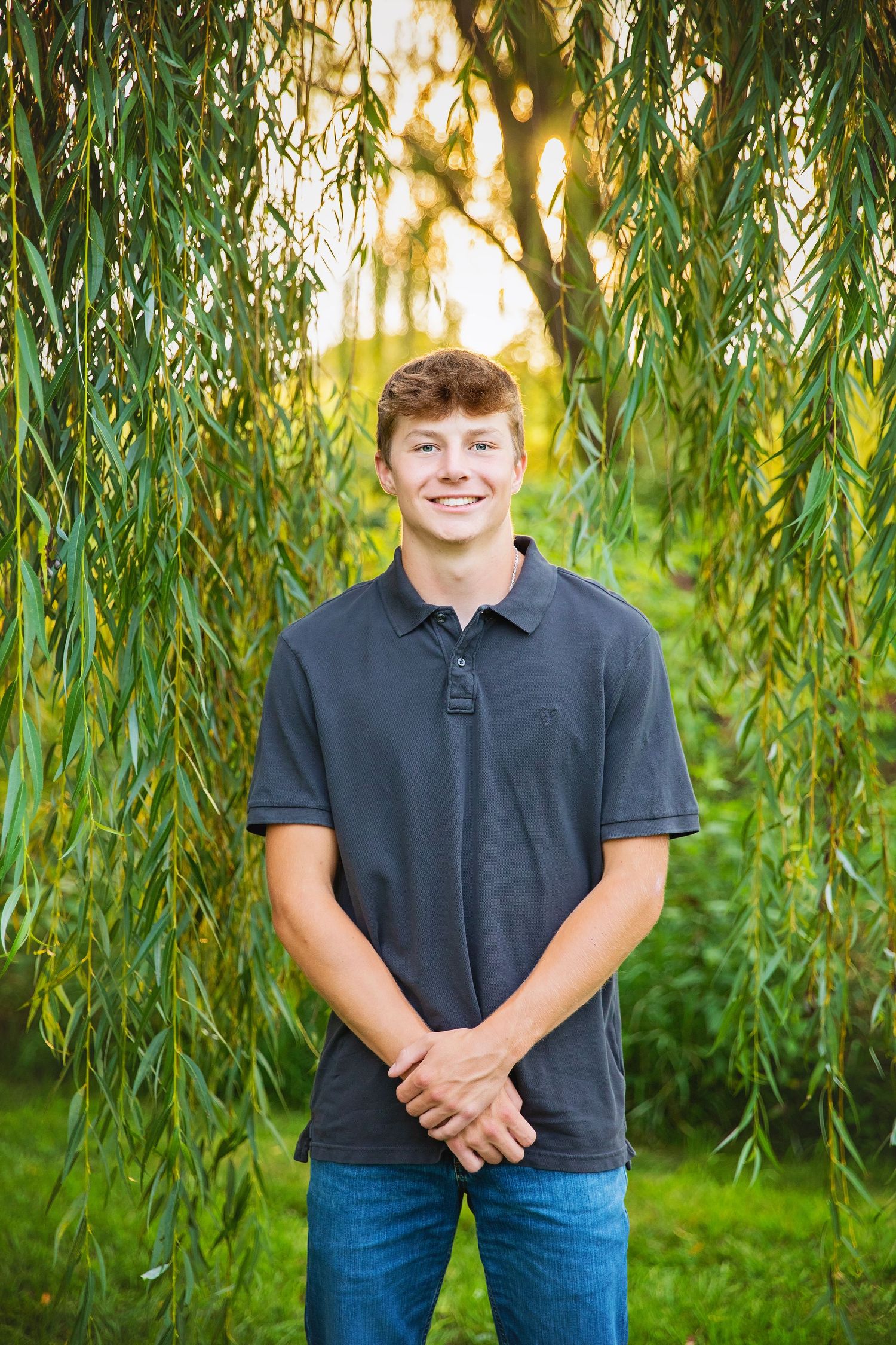 Smiling teenage boy standing under weeping willow tree branches in a dark polo shirt.