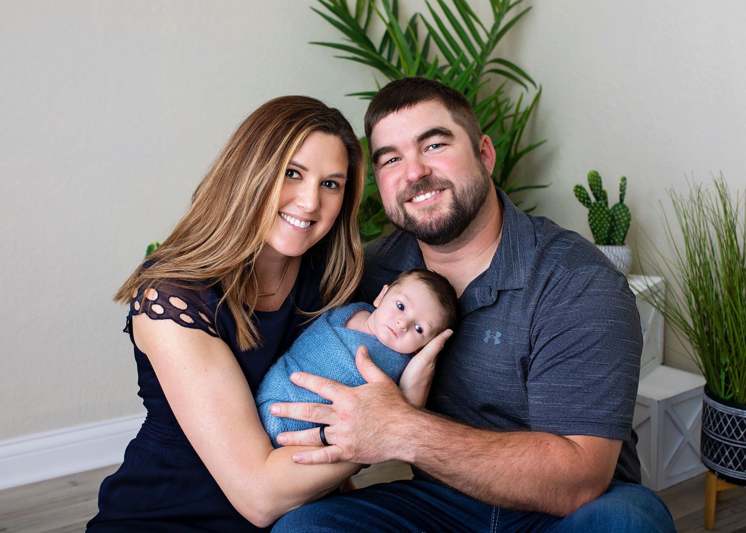 Smiling parents sitting together holding their newborn baby boy wrapped in a blue swaddle.