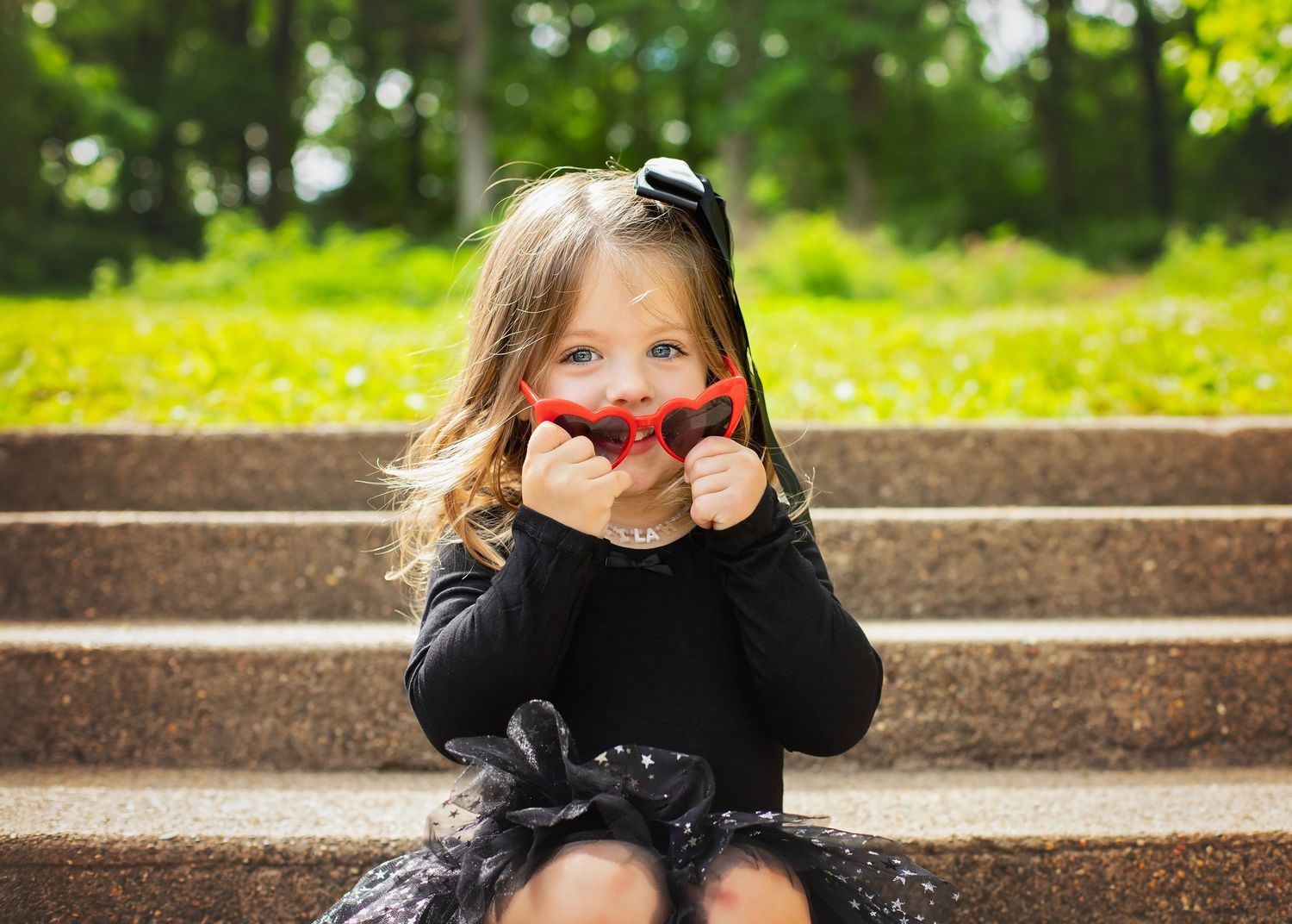 Adorable little girl in black outfit holding red heart sunglasses while sitting on stone steps outdoors.