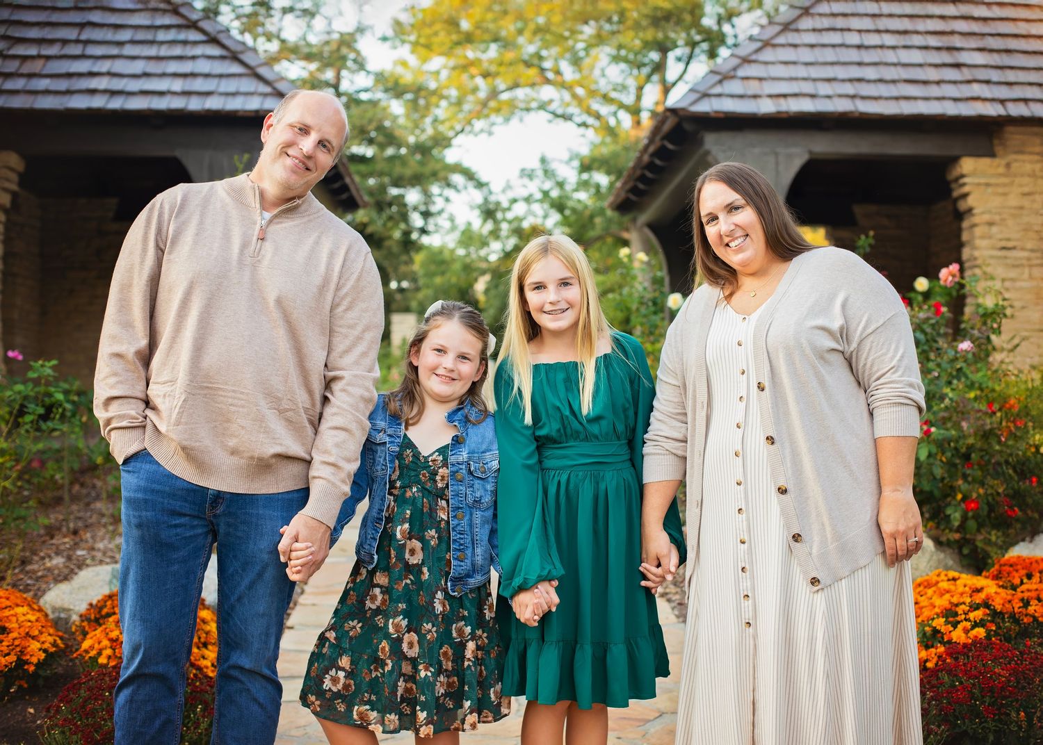 Family of four holding hands and smiling outdoors near a building surrounded by autumn flowers and foliage.