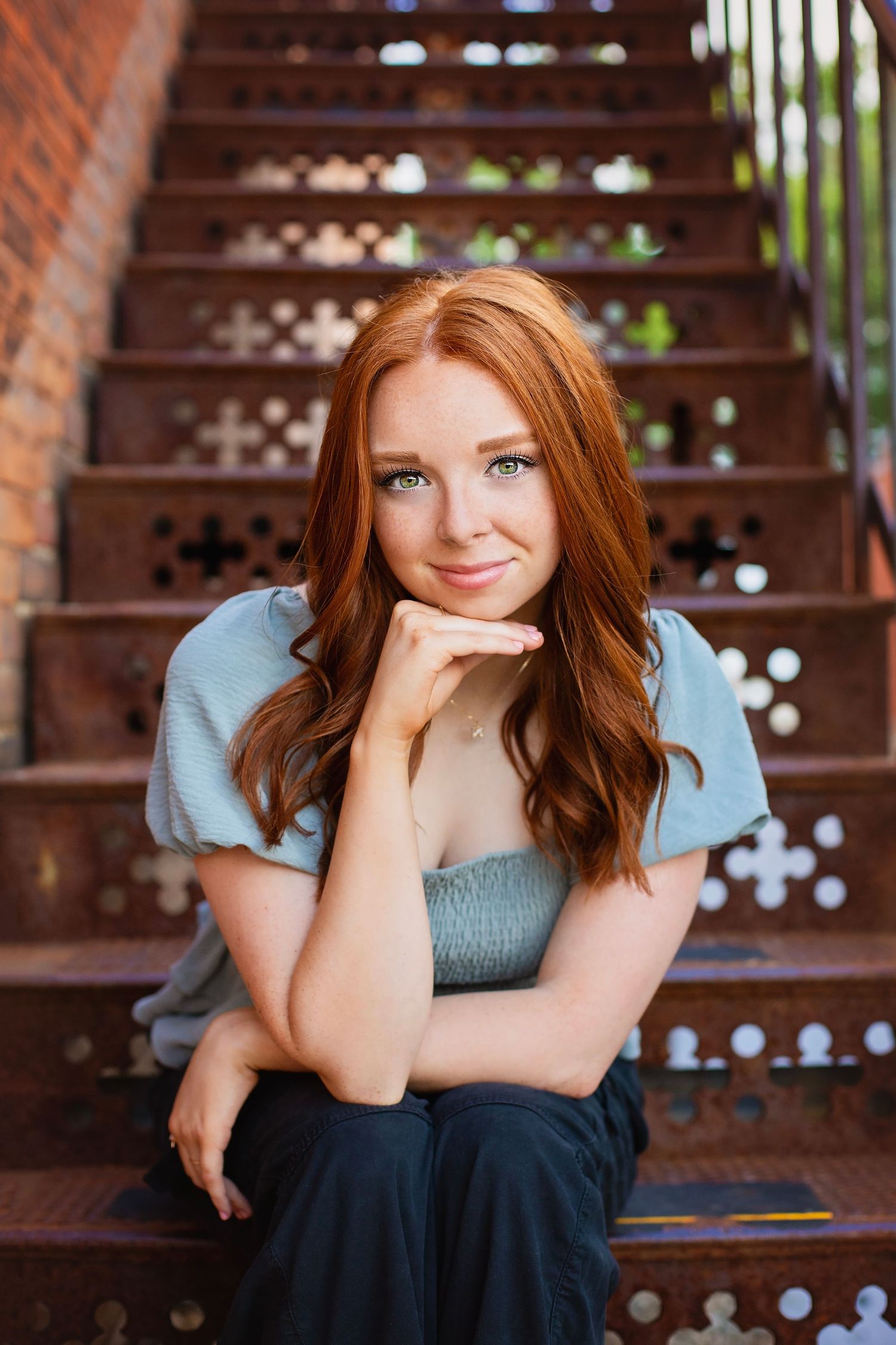 Young redhead woman with freckles sits on ornate metal stairs, resting chin on hand, smiling softly at camera.