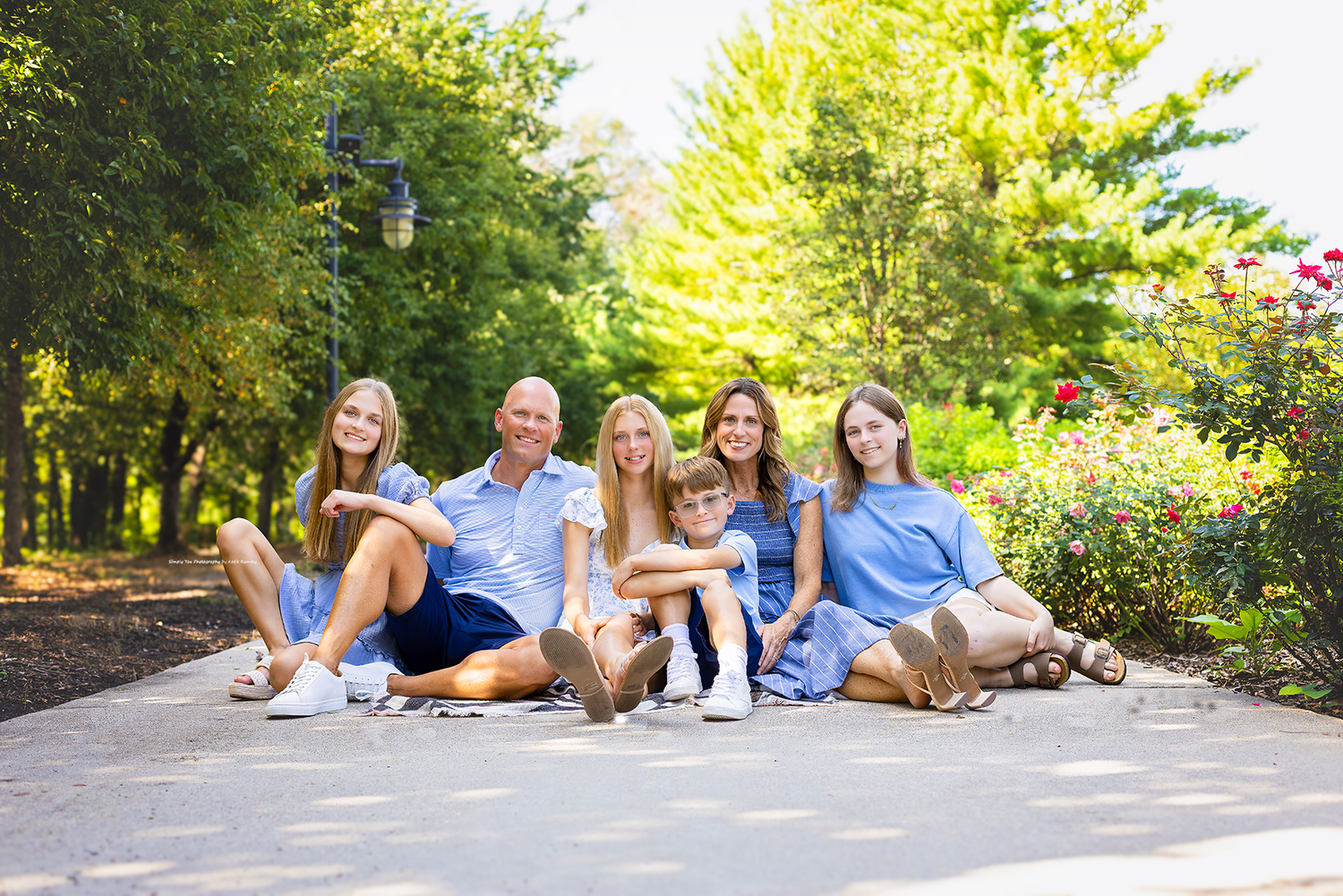 Happy family of six sitting together on a garden path surrounded by lush green trees and colorful flowers.