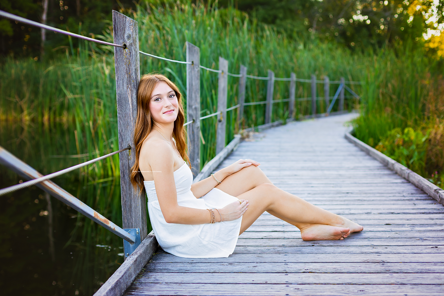 Young woman in white dress sitting on wooden bridge boardwalk surrounded by lush green reeds near water.