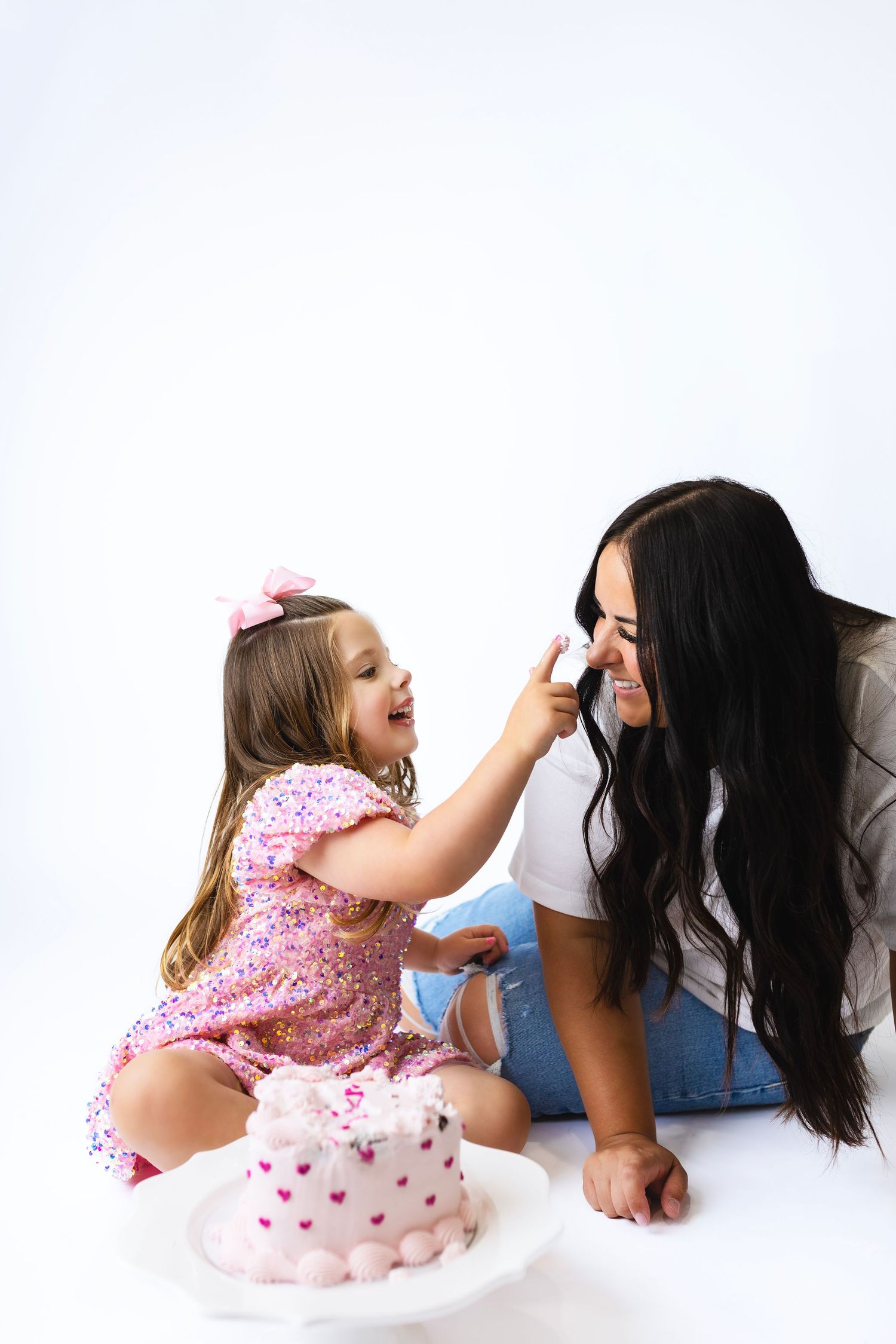 A laughing toddler in a pink dress playfully puts frosting on her mom's nose beside a heart-decorated birthday cake.