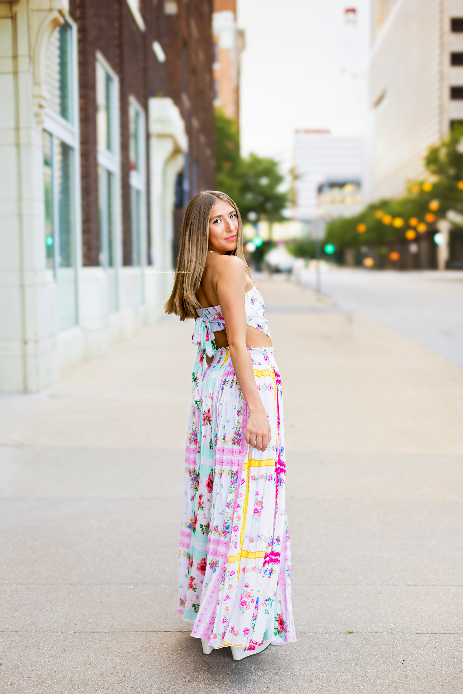 Young woman in colorful floral maxi dress posing on urban sidewalk, looking over shoulder.