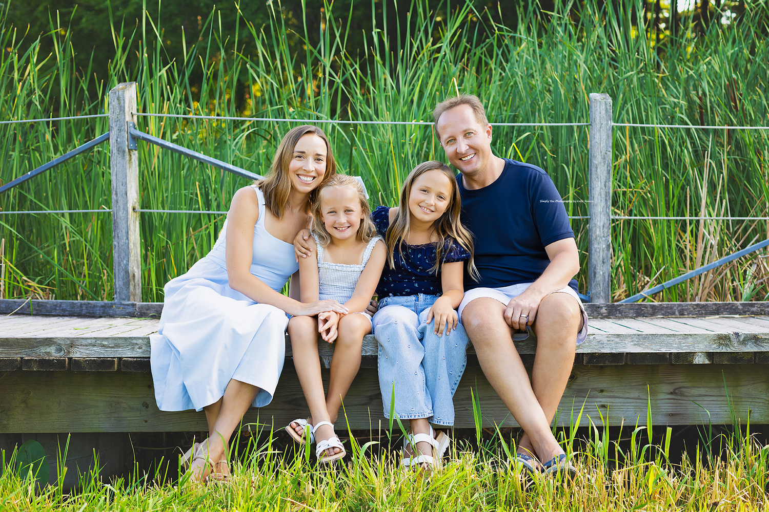 Happy family of four sitting on a wooden dock surrounded by lush green reeds on a sunny day.