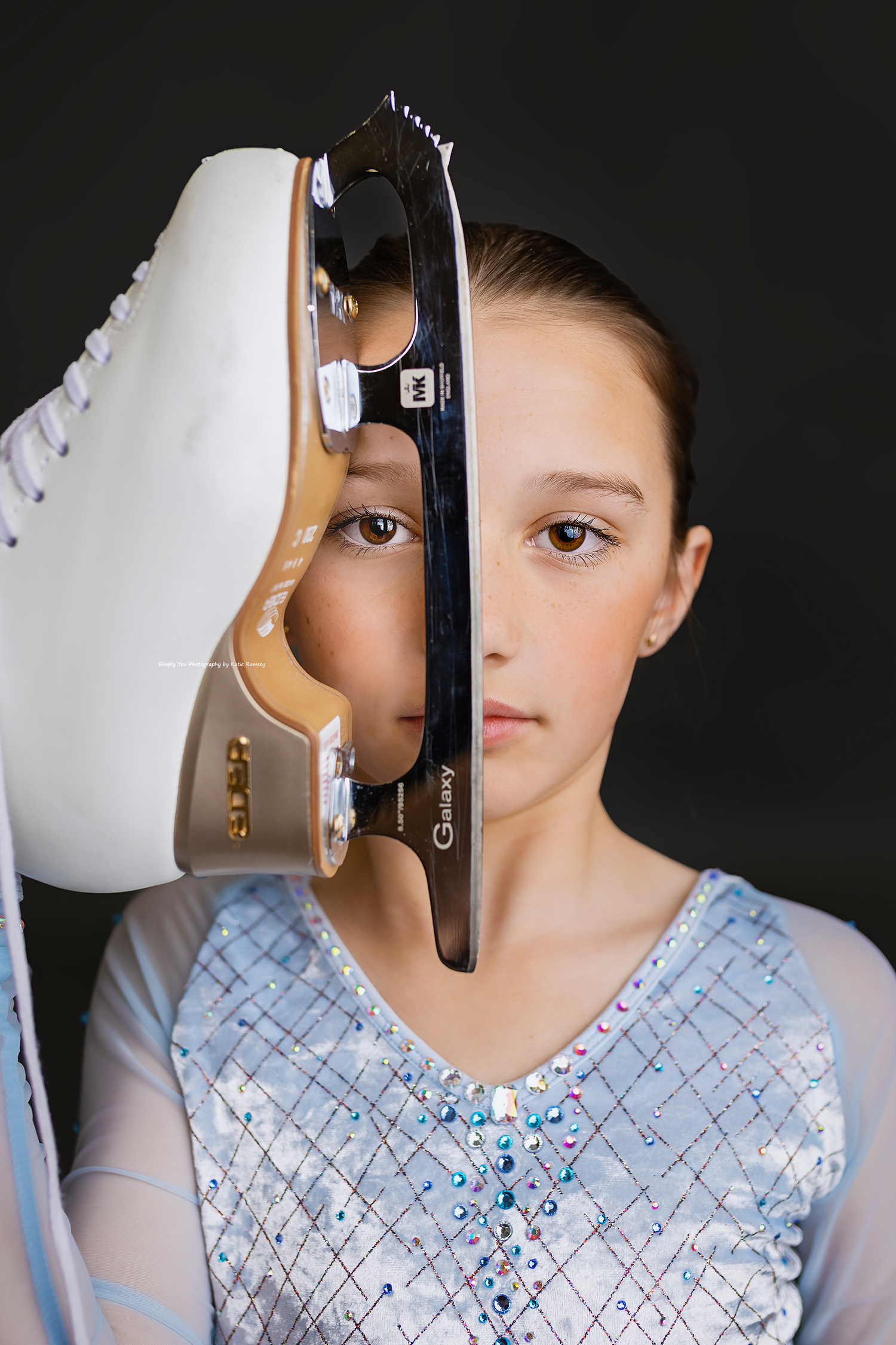 Young figure skater in blue rhinestone dress holds ice skate blade up to her face against dark background.