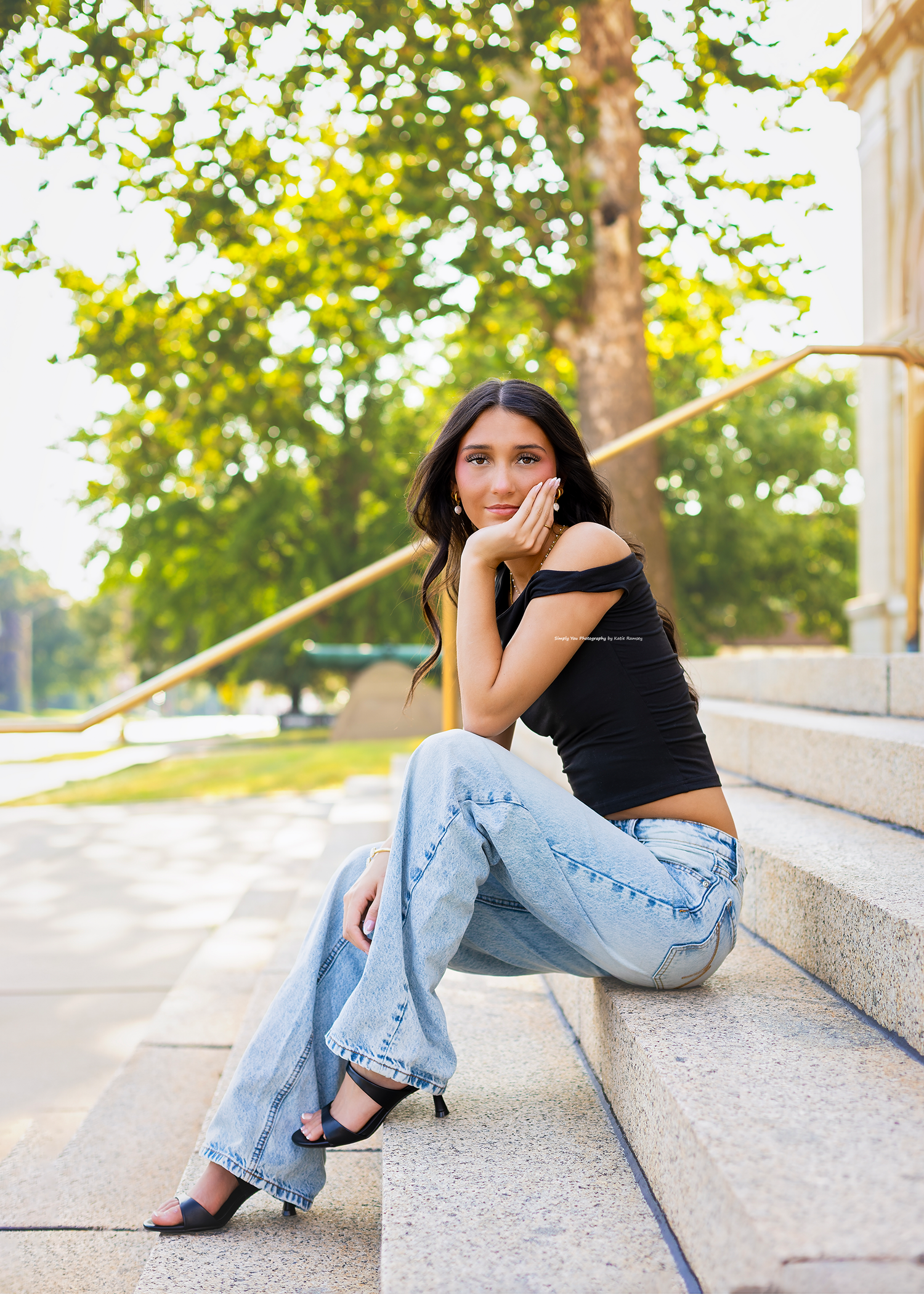Young woman in black crop top and wide-leg jeans sits on stone steps outdoors, posing with hand on chin.