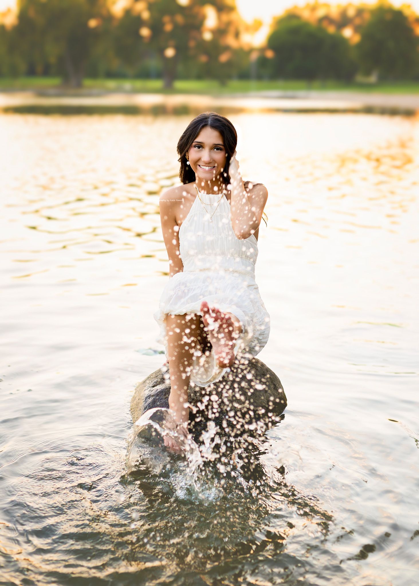 Joyful young woman in white dress splashing water while sitting on rock in sunlit lake.