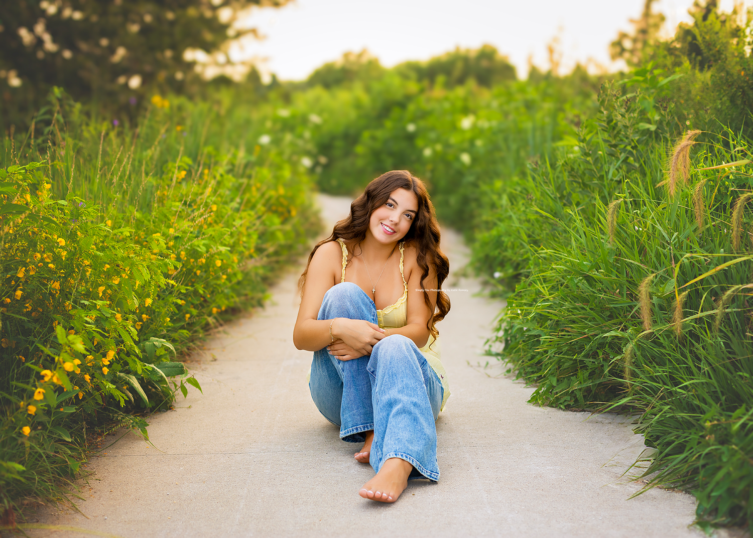 Young woman in yellow top and jeans crouching on a path surrounded by lush green plants and yellow wildflowers.