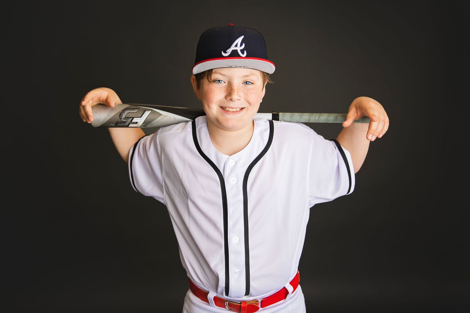 Smiling youth baseball player in Braves uniform resting bat across shoulders, dark studio background.