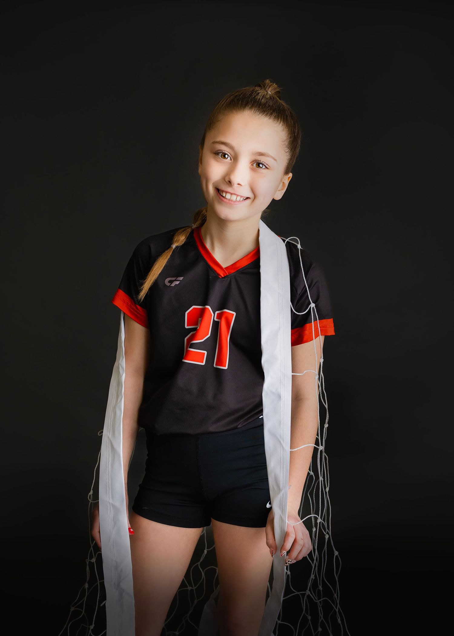 Smiling young girl in number 21 black volleyball jersey stands holding a volleyball net on a dark studio background.