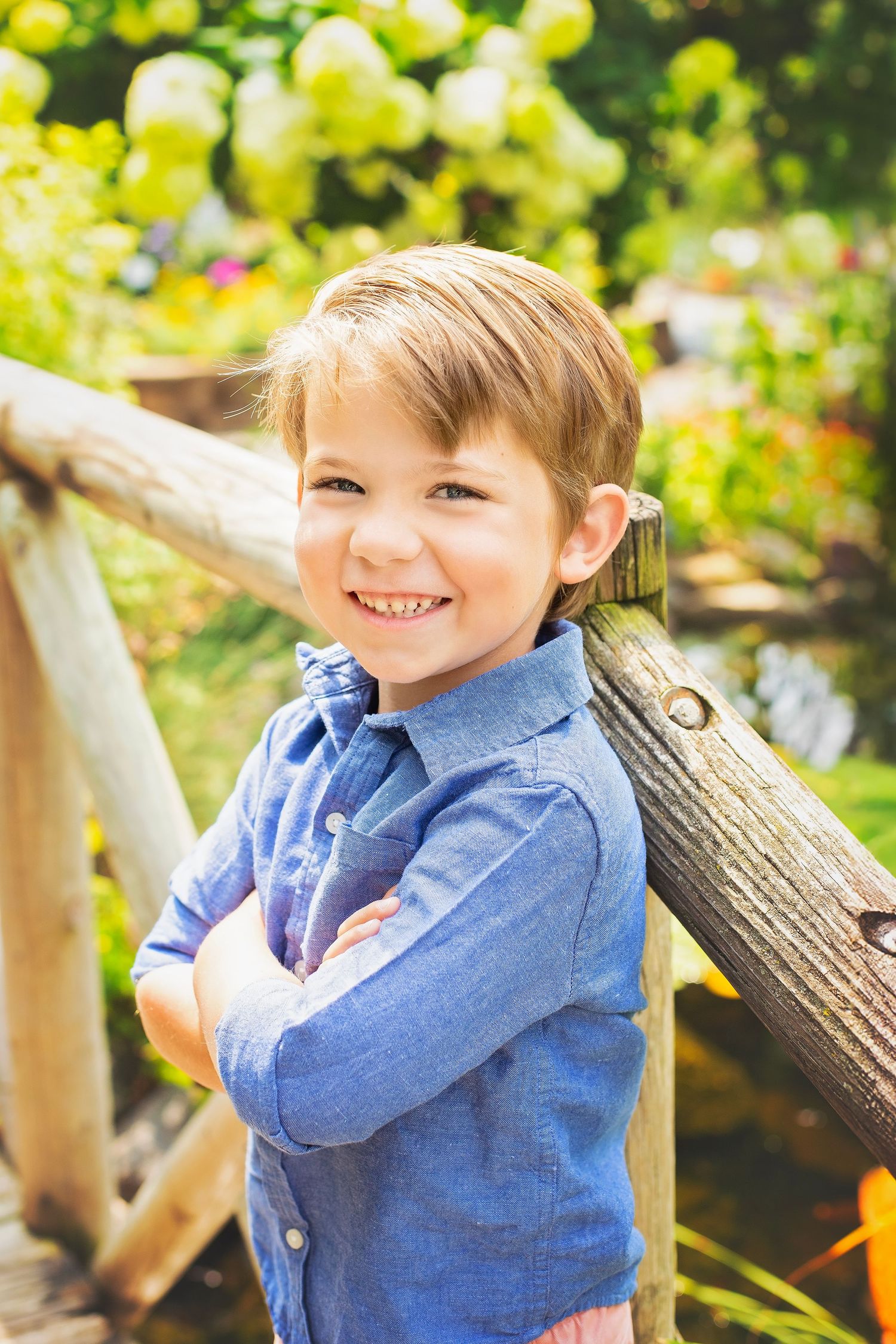 Cheerful young boy in blue shirt stands with crossed arms smiling on sunny garden bridge.