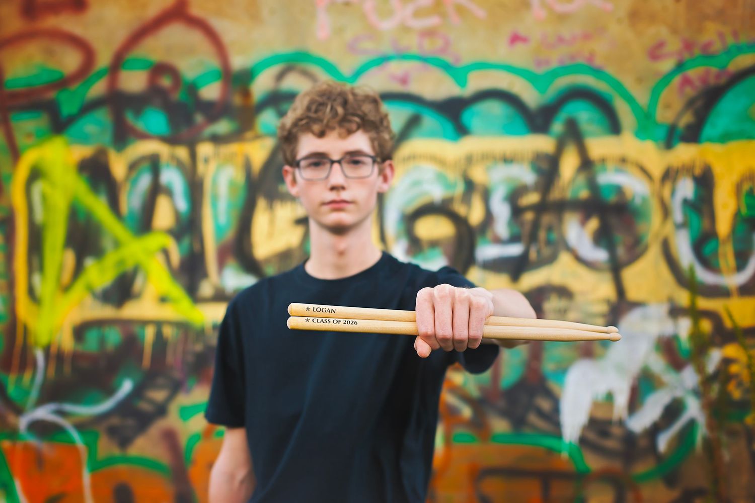 A teenage boy with glasses holds drumsticks toward the camera in front of a colorful graffiti wall.