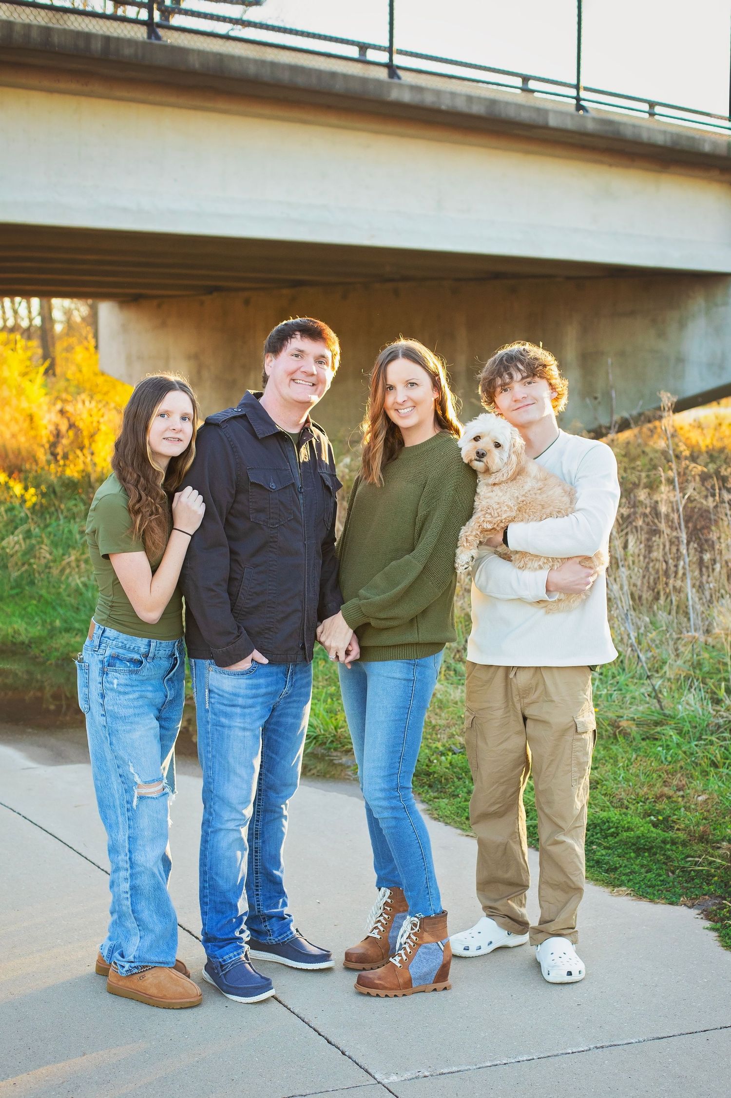 Family of four with a small dog posing near a bridge in fall.
