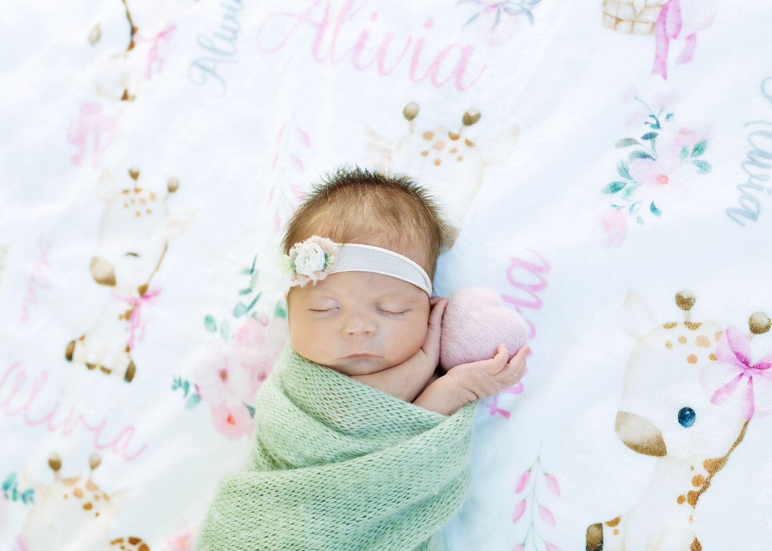 Sleeping newborn baby girl swaddled in green wrap lying on personalized floral name blanket.