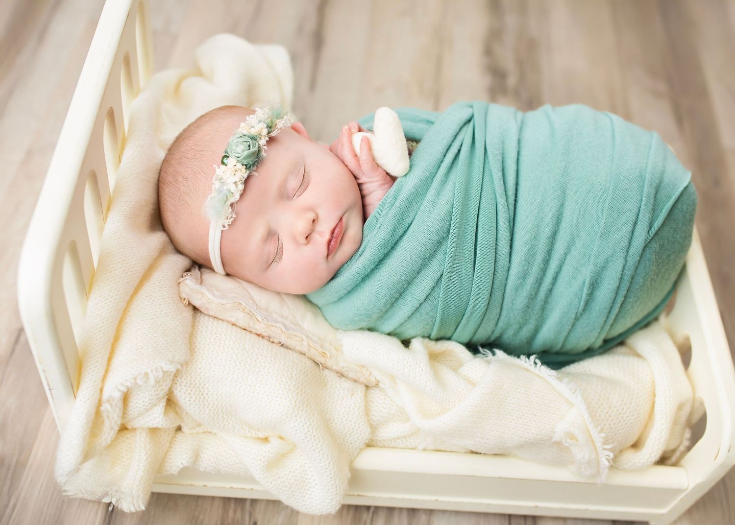 Sleeping newborn swaddled in teal wrap with floral headband resting in small white wooden crib.
