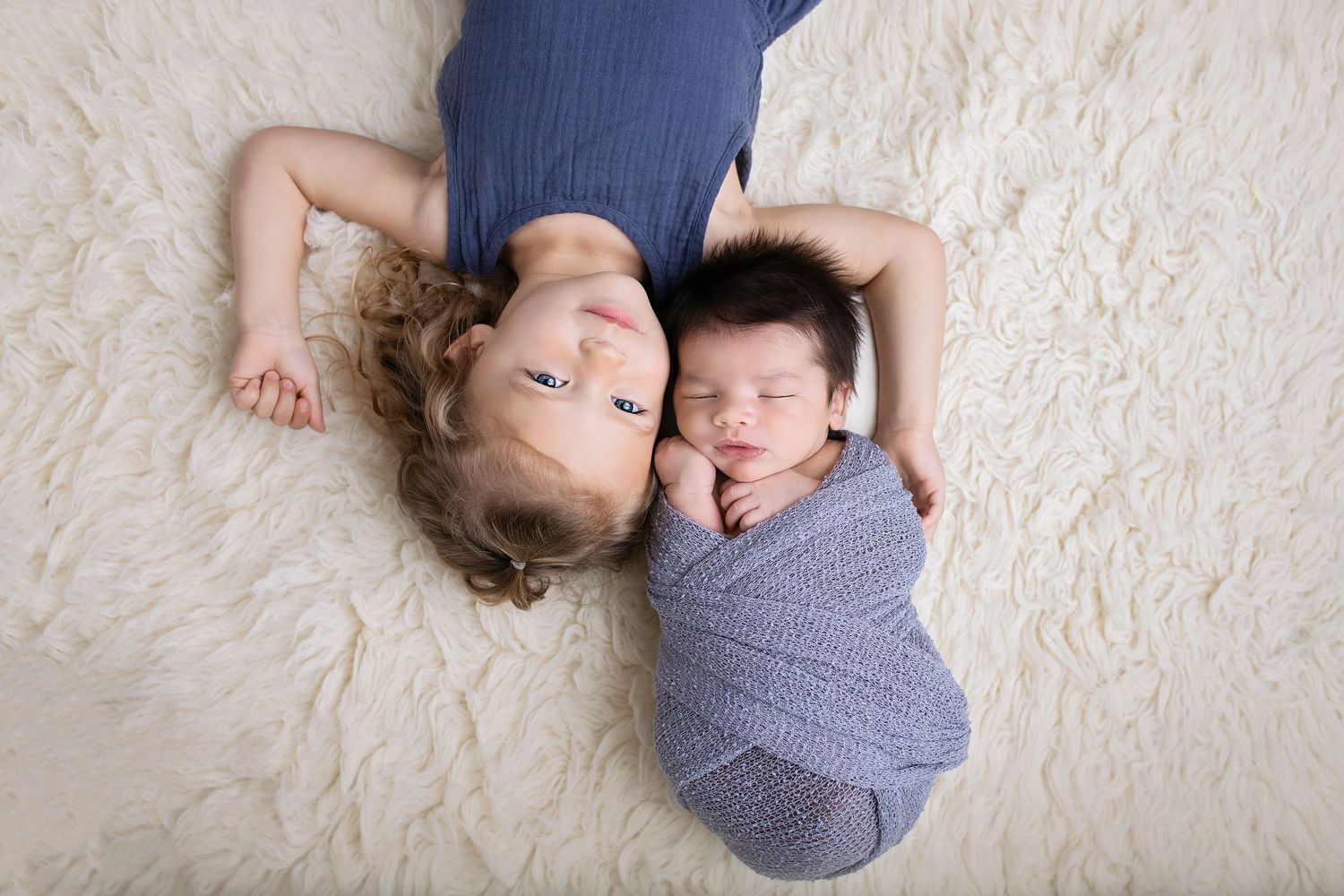 Young child lying next to sleeping newborn baby wrapped in blue swaddle on white rug.