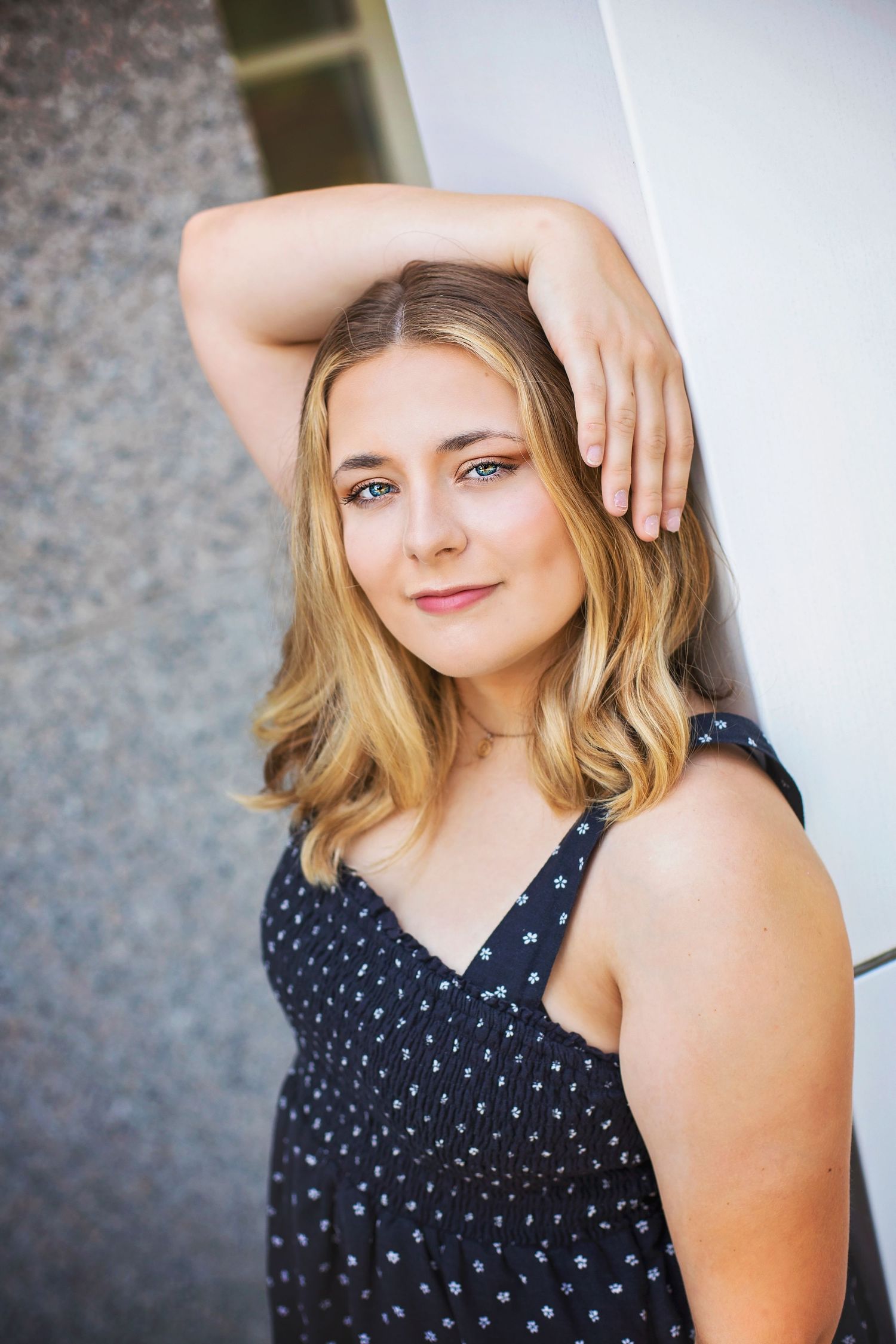 Young blonde woman in navy polka dot dress leaning against white wall with hand raised to head.