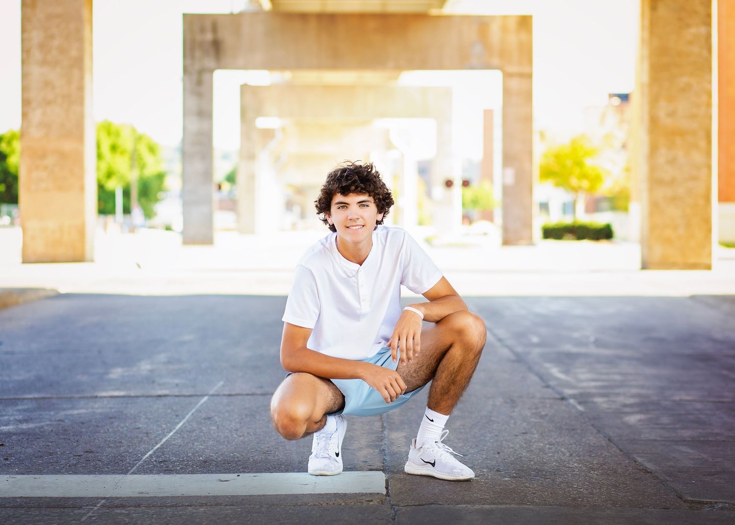 Smiling teen boy in white outfit crouches on a paved plaza under concrete archways outdoors.