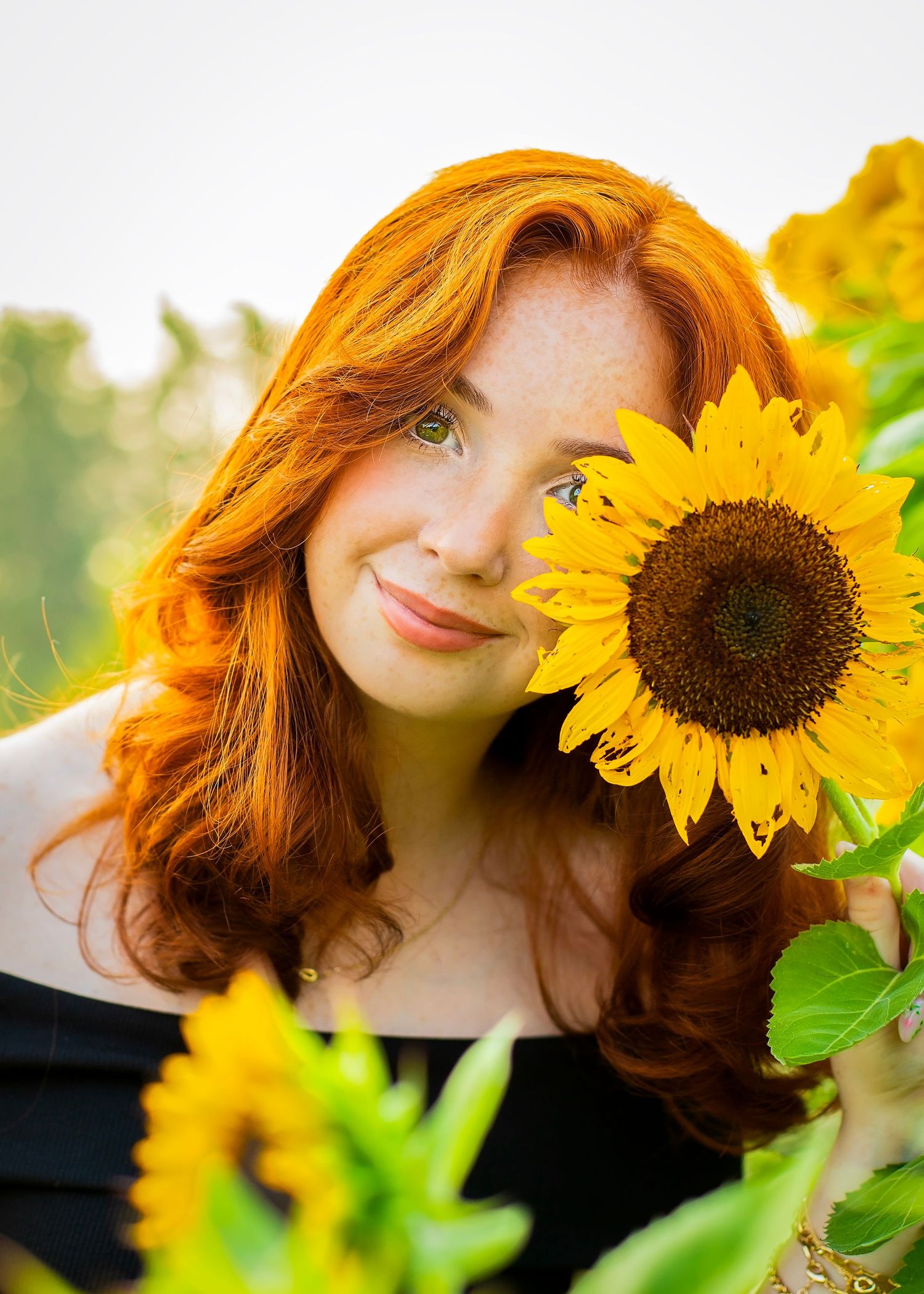 Beautiful redhead girl with freckles posing with a bright yellow sunflower outdoors.