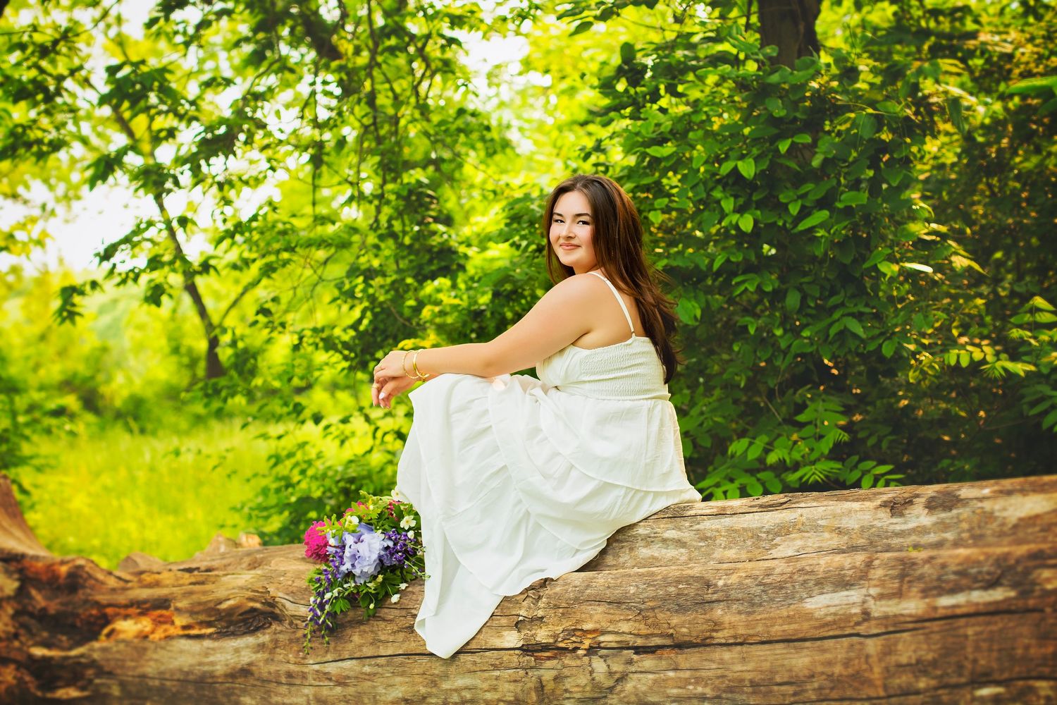Young woman in white dress sitting on fallen log surrounded by green trees.