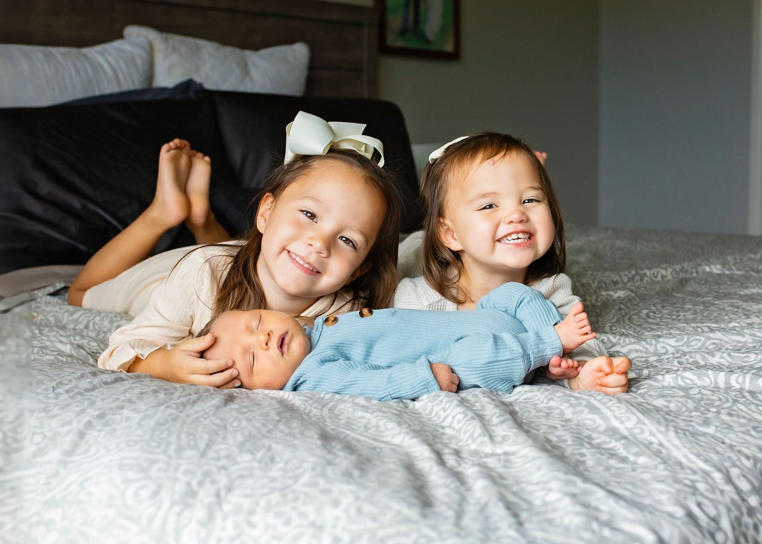 Two smiling sisters lying on a gray bed with their newborn baby brother dressed in blue between them.