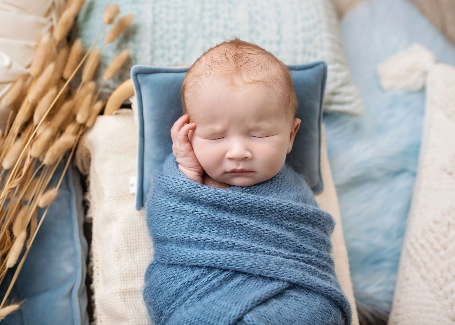 Peaceful newborn baby wrapped in a blue knit blanket resting on a tiny bed with dried floral decor.