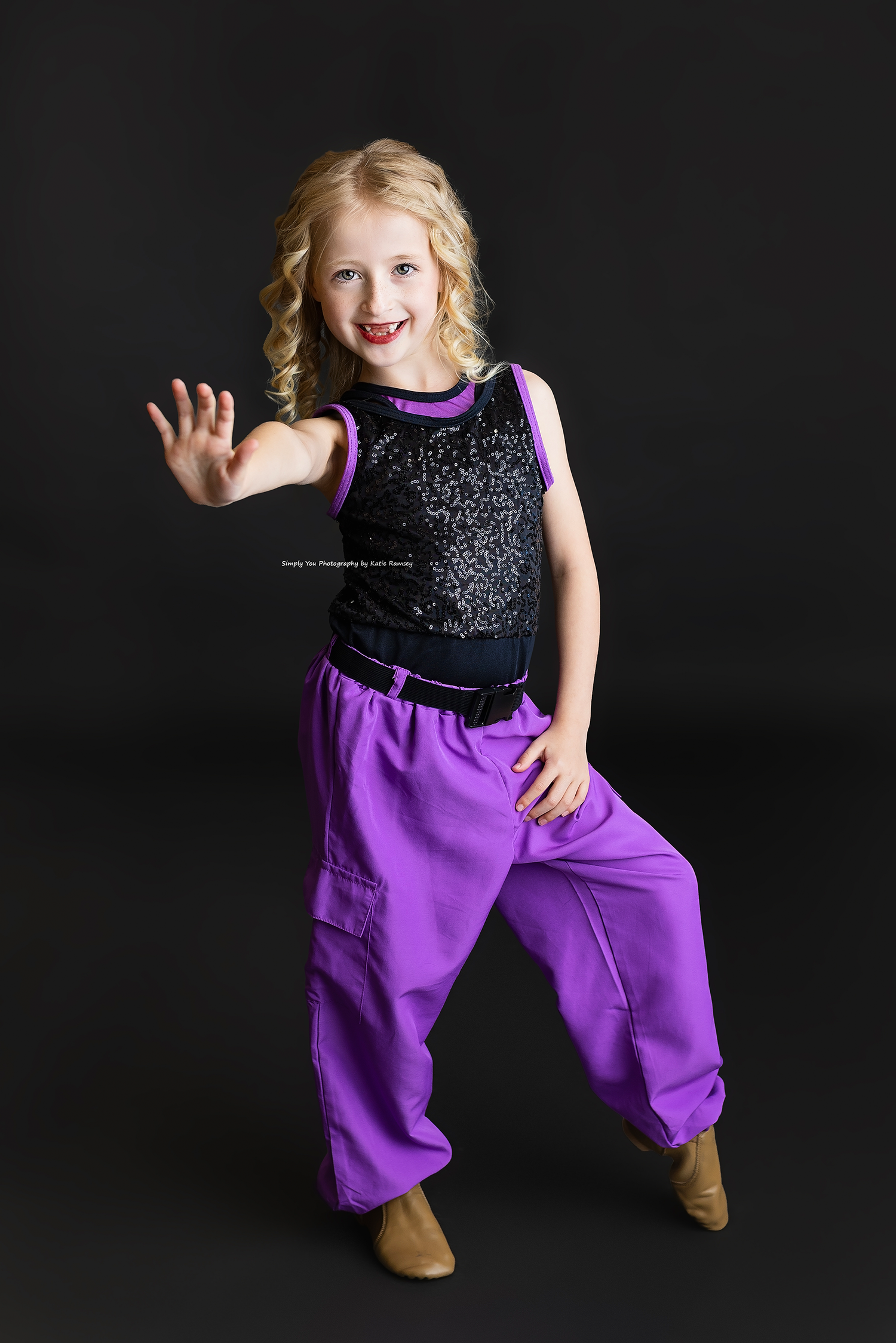 Young blonde girl in purple harem pants and black sequin top poses playfully against dark background.